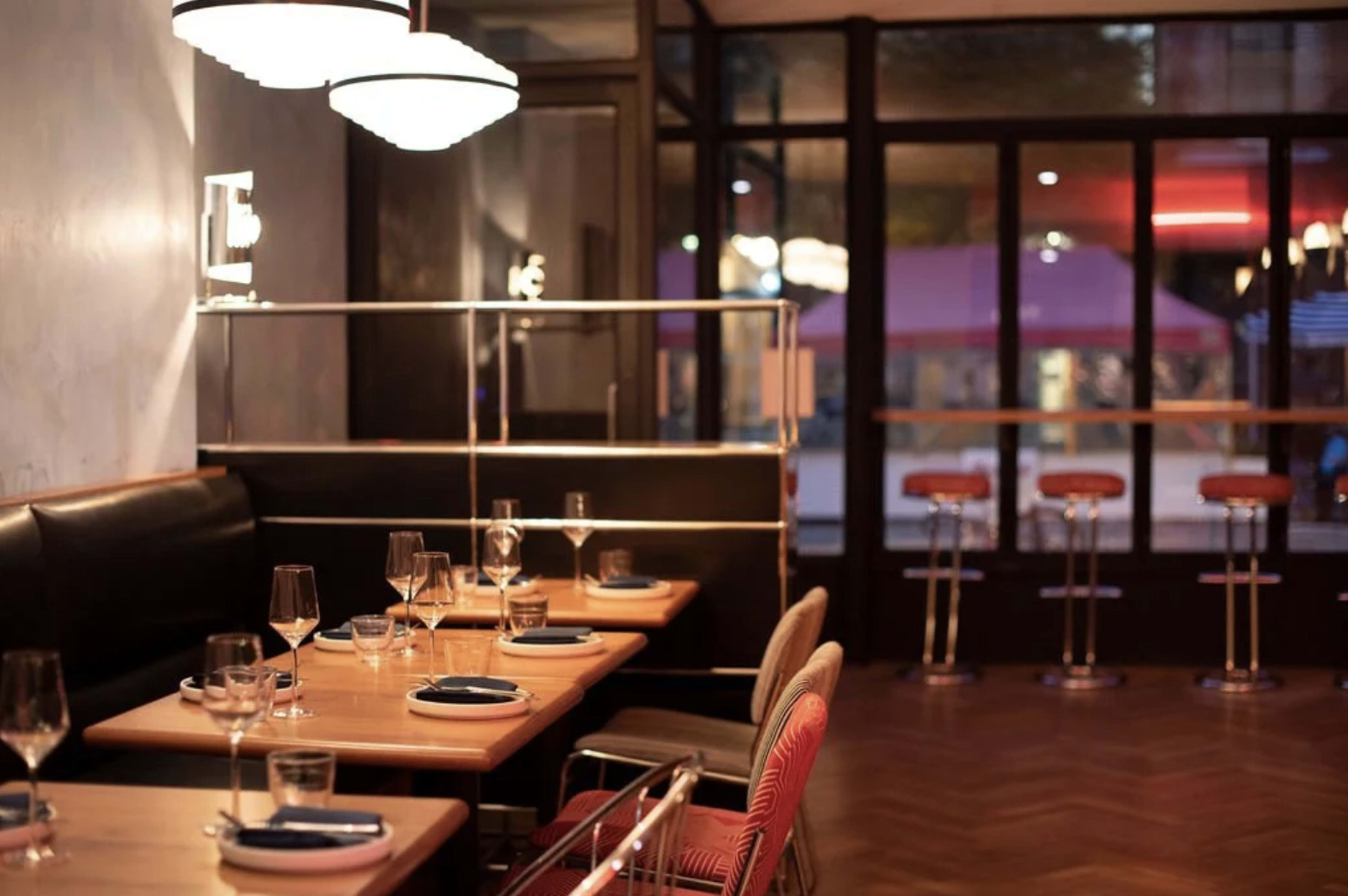 The image shows a modern restaurant interior with wooden tables set for dining, stylish lighting fixtures, and bar stools visible near a large window.