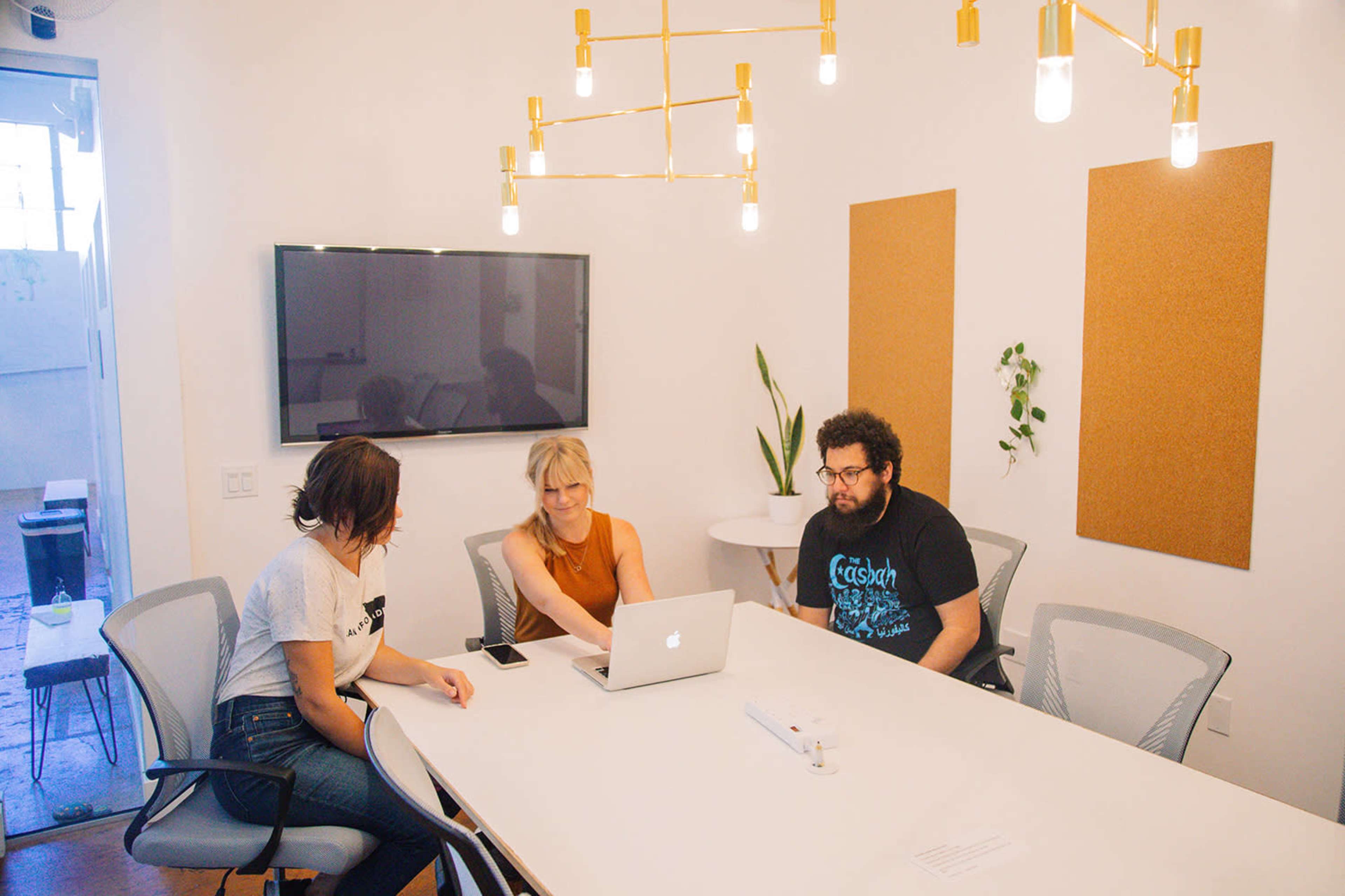 Three people sit at a modern conference table working on a laptop in a well-lit meeting room.