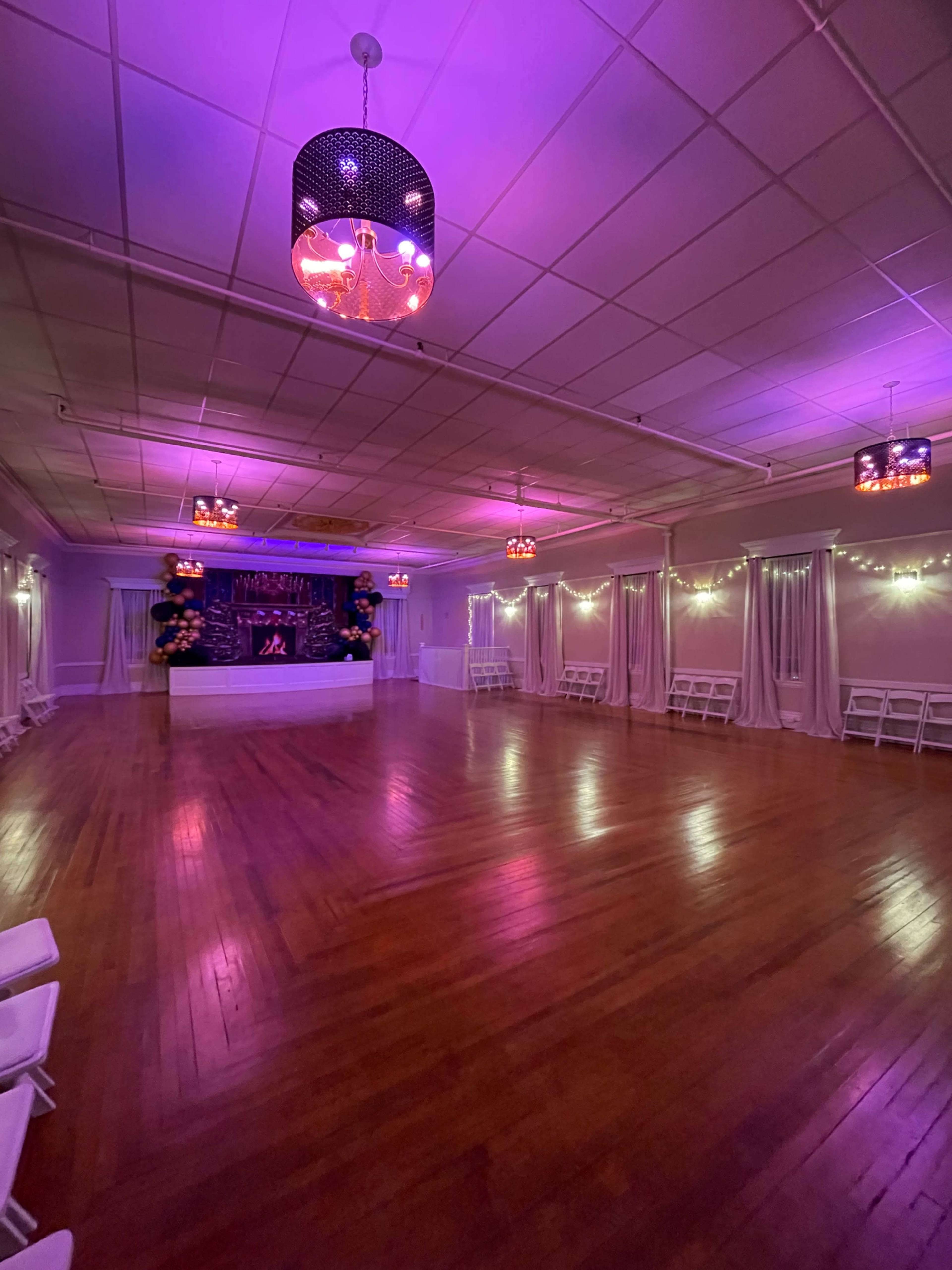 The image shows an empty event space with wooden flooring, purple lighting, and white folding chairs arranged along the sides.