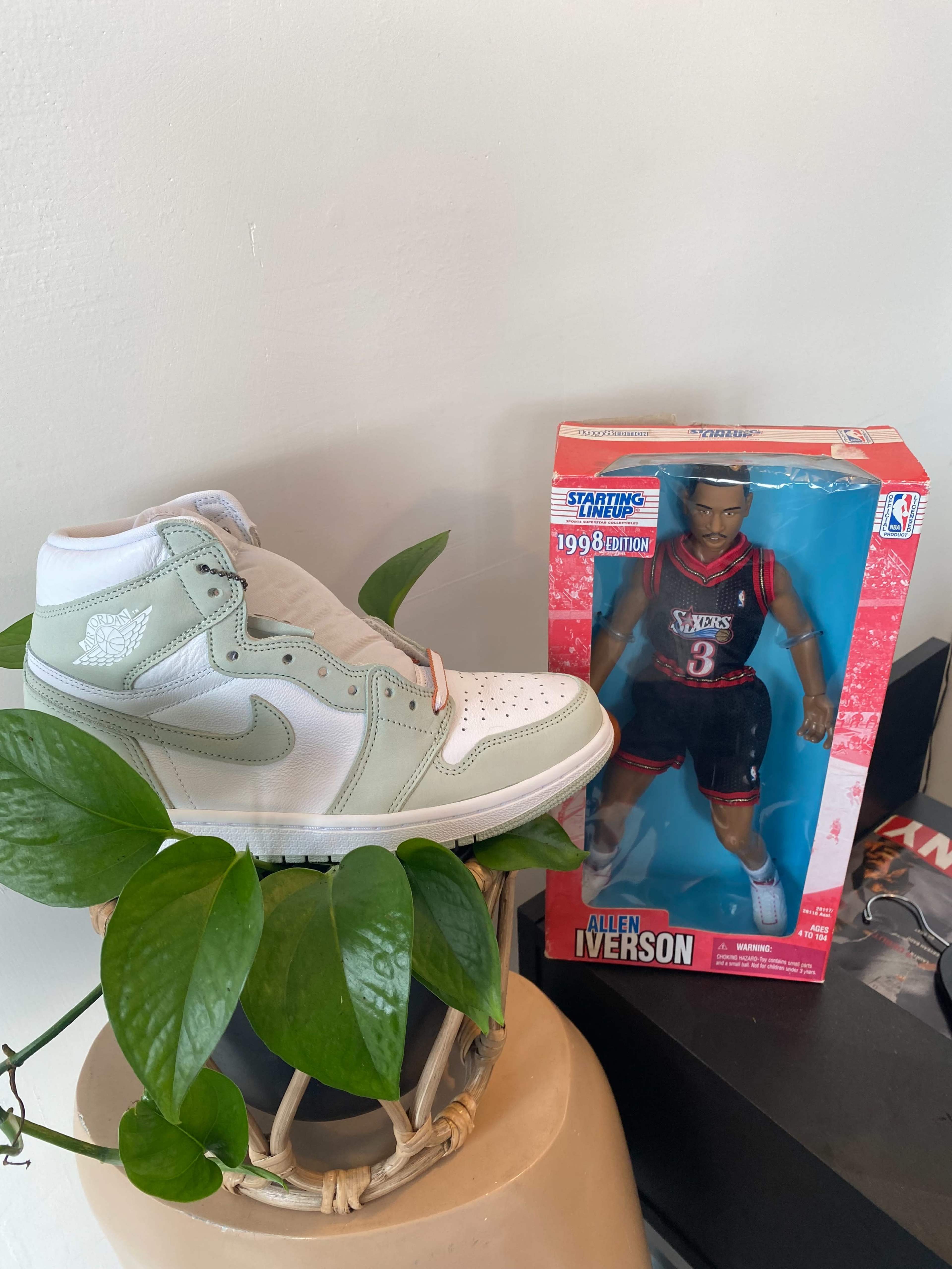 A pair of white and mint green sneakers sits next to a boxed action figure of Allen Iverson wearing a red basketball jersey.