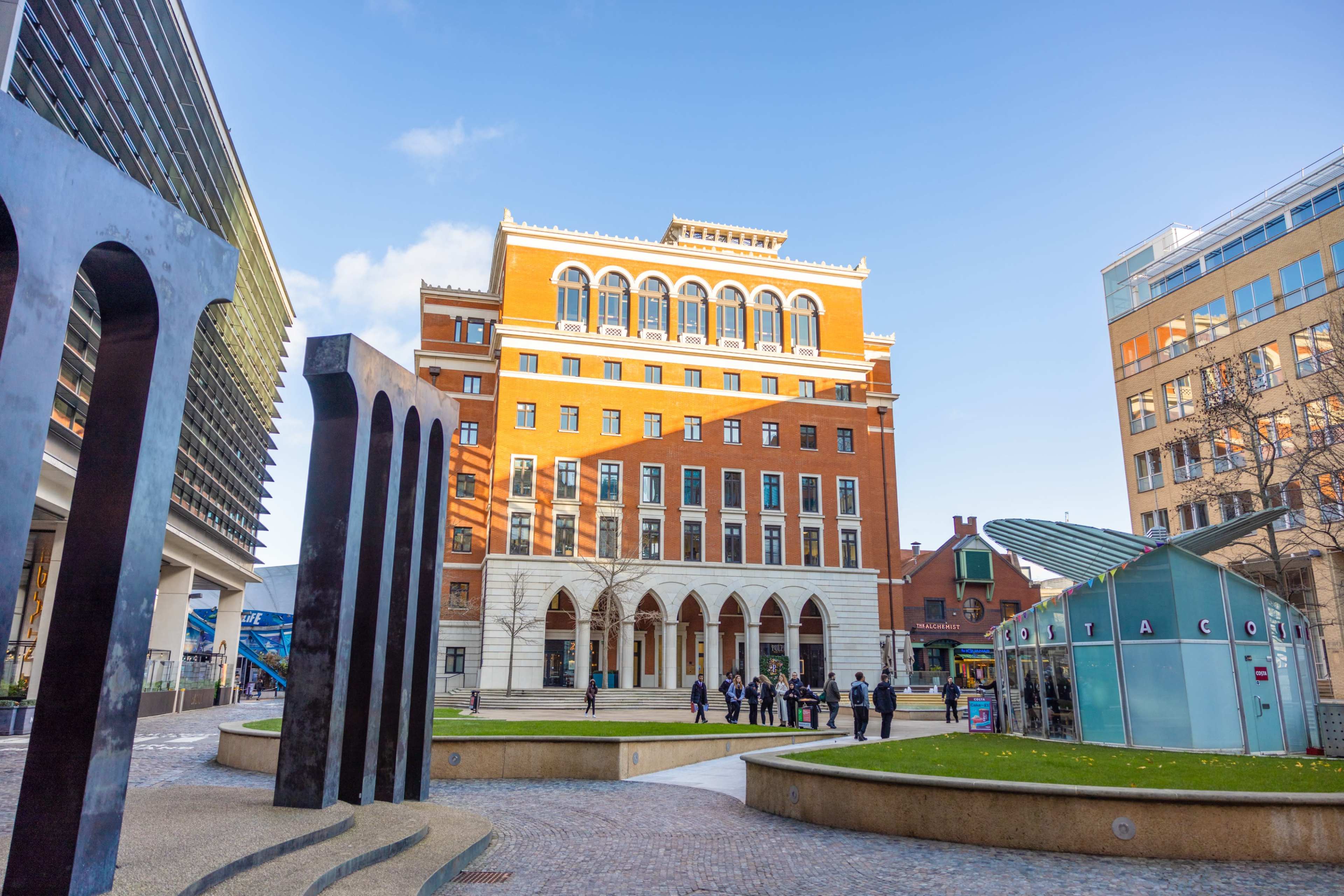 A spacious urban plaza featuring modern sculptures, a historic brick building with arched windows, and groups of people gathered in the open area.