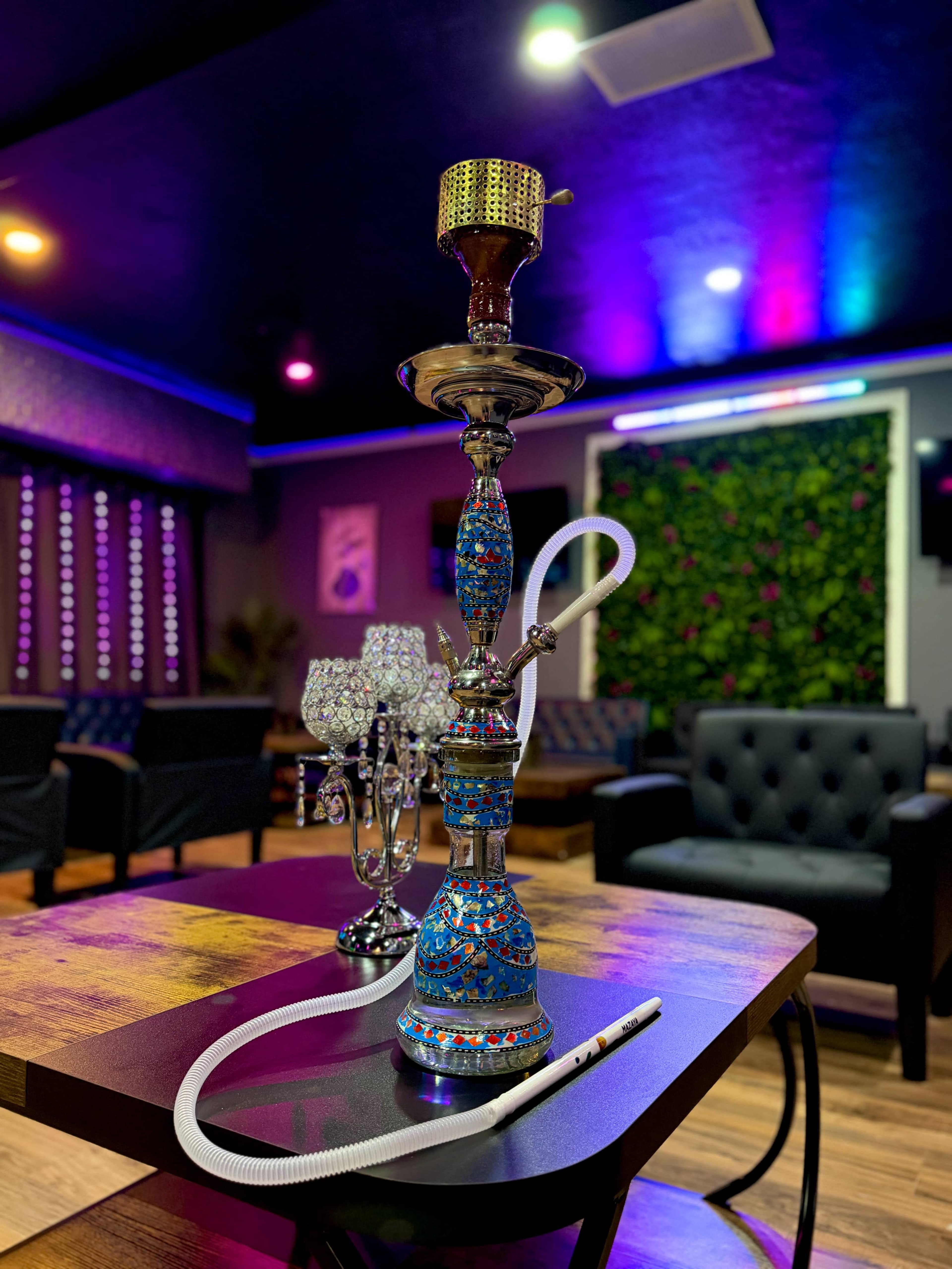 A decorated hookah stands on a wooden table in a dimly lit lounge adorned with colorful lights and plush seating.