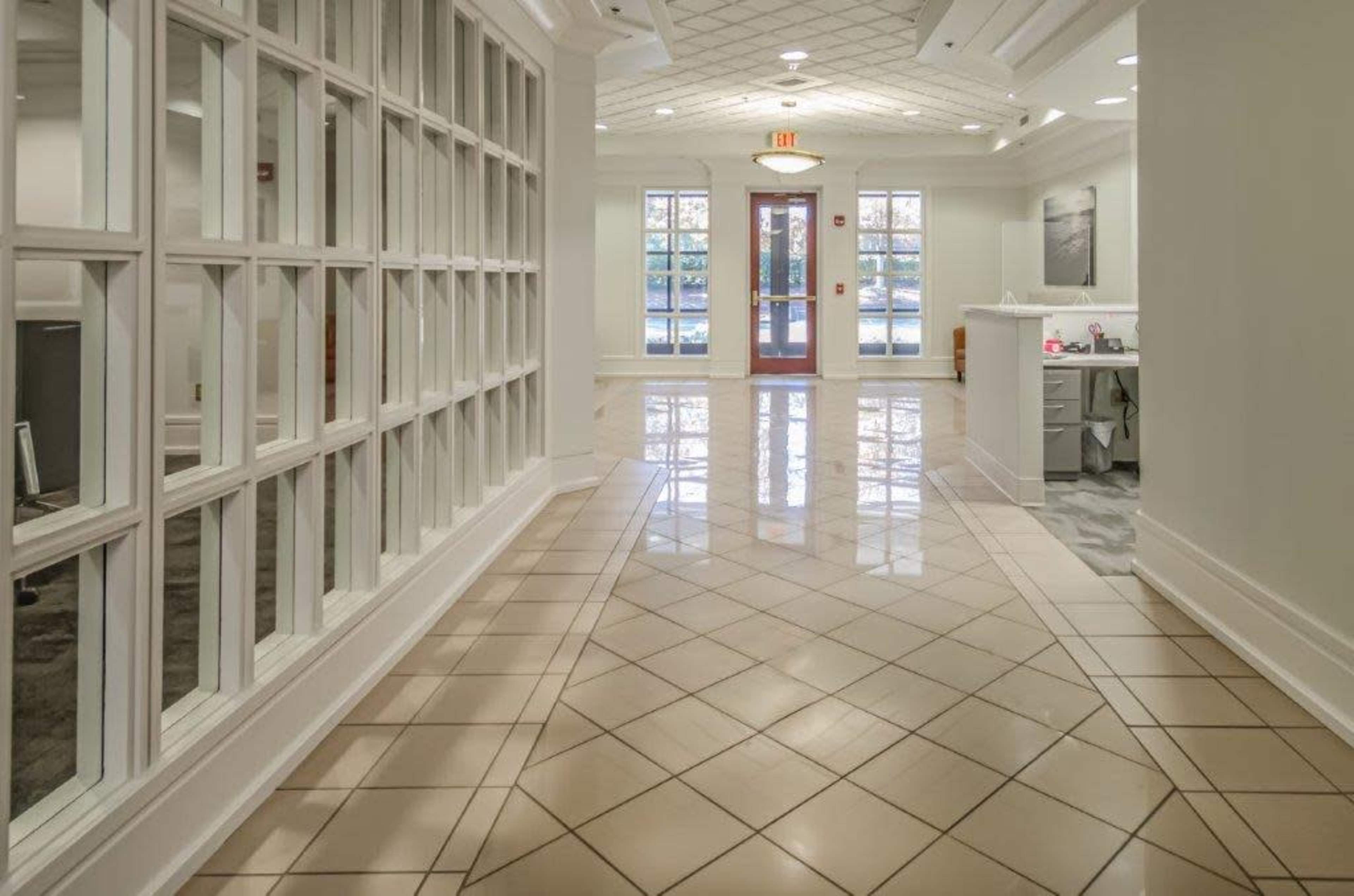 A hallway with large windows on one side leads to a reception area, featuring polished tile floors and a bright entrance.