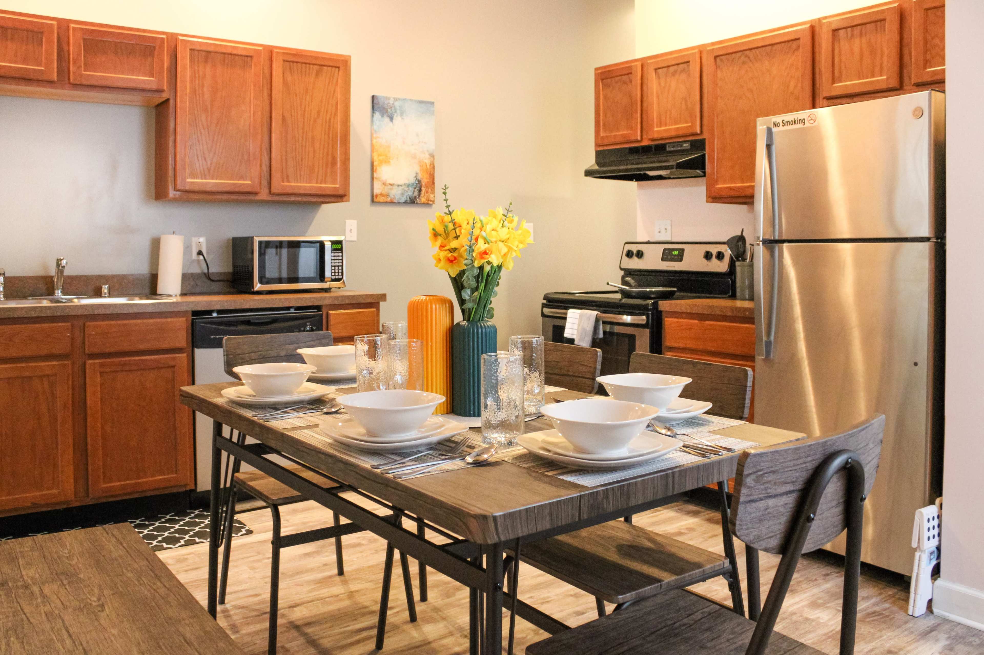 A modern kitchen features wooden cabinets, stainless steel appliances, and a dining table set for six with bowls and glasses, complemented by a floral centerpiece.