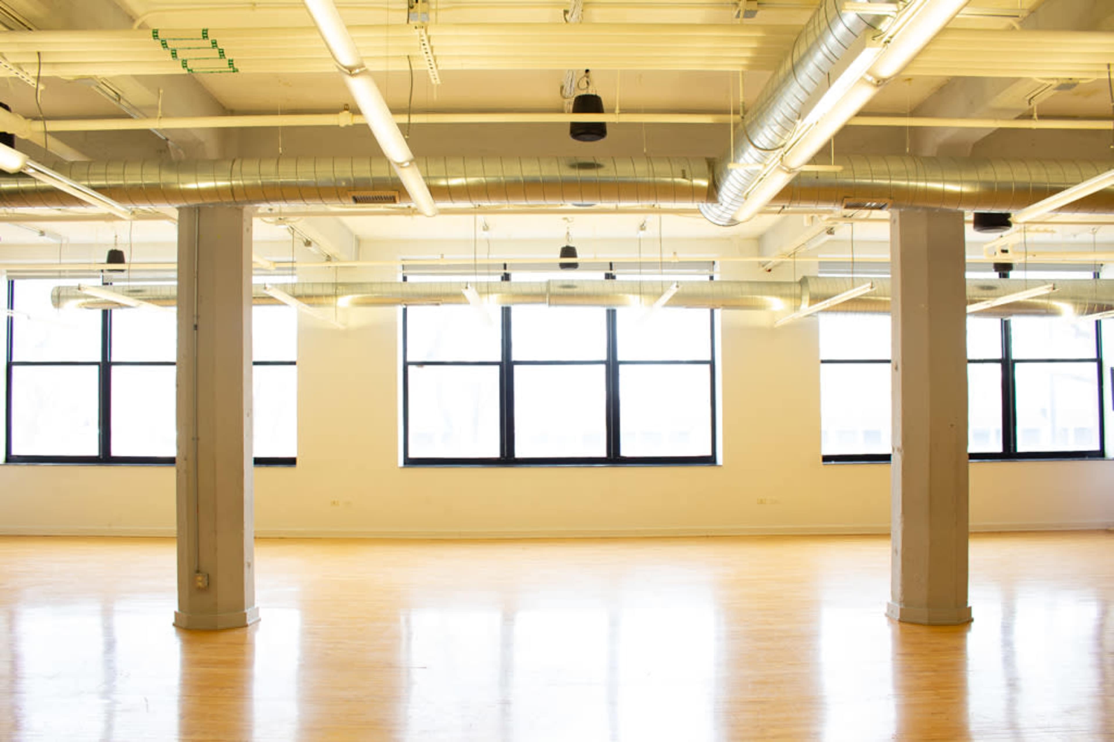The image shows an empty room with wooden flooring, large windows, and exposed ductwork on the ceiling.