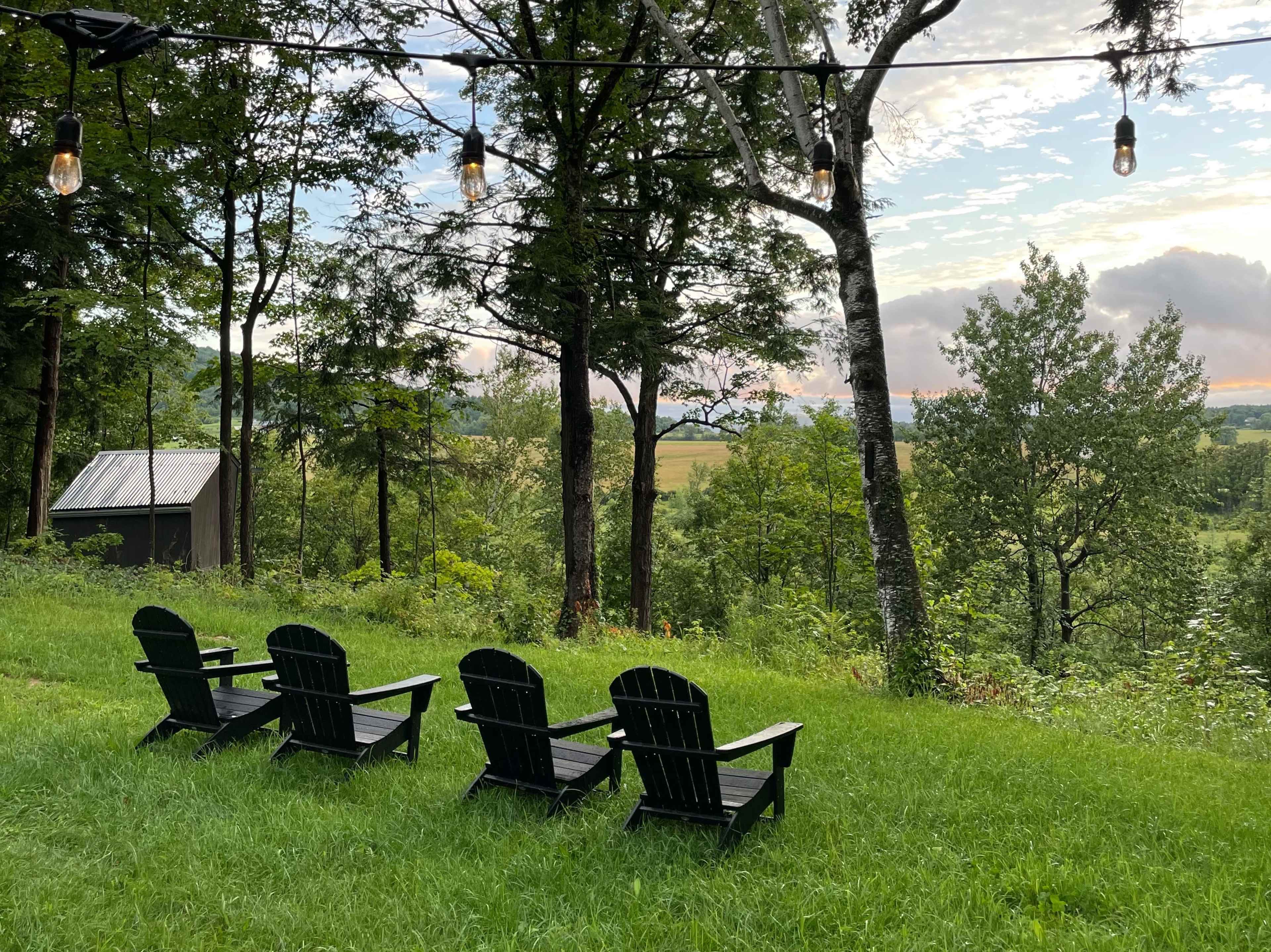 Four black adirondack chairs are positioned on a grassy area overlooking a scenic landscape with trees and an open field.