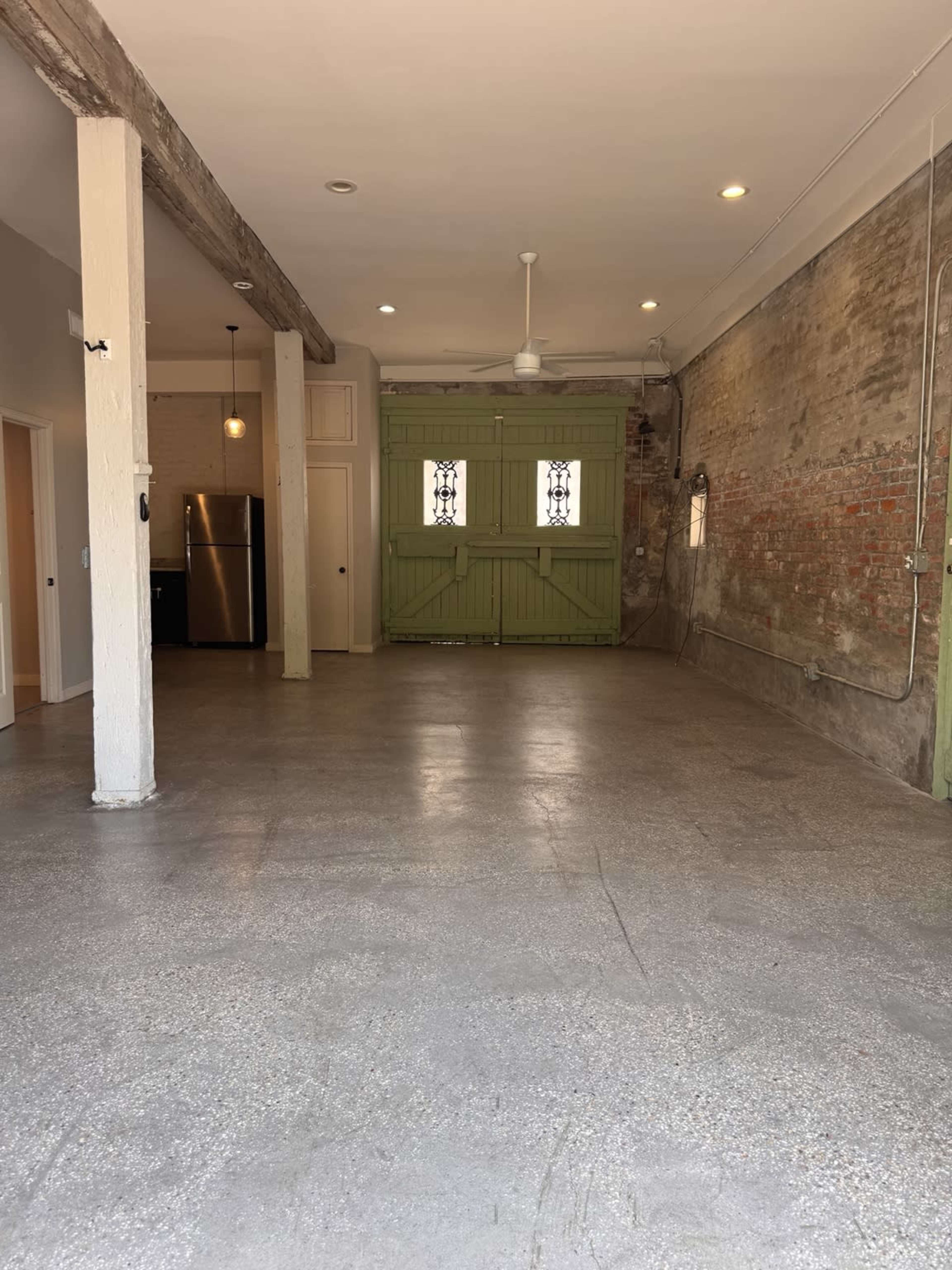 The image shows a spacious, empty room with a concrete floor, exposed brick walls, and a green double door at one end.