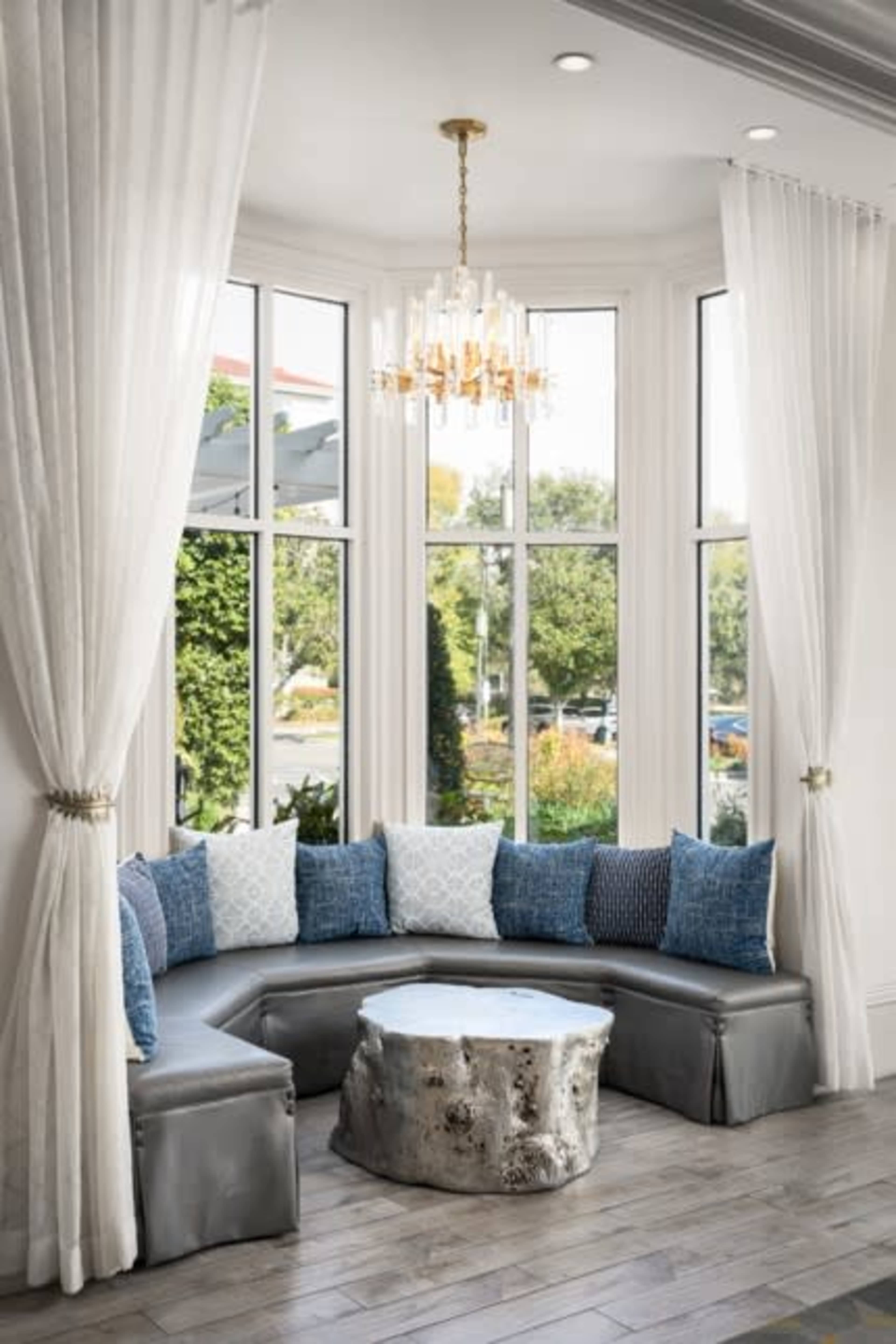A curved seating area with gray cushions and blue decorative pillows is placed in front of large windows, accompanied by a round, natural wood coffee table and a chandelier above.