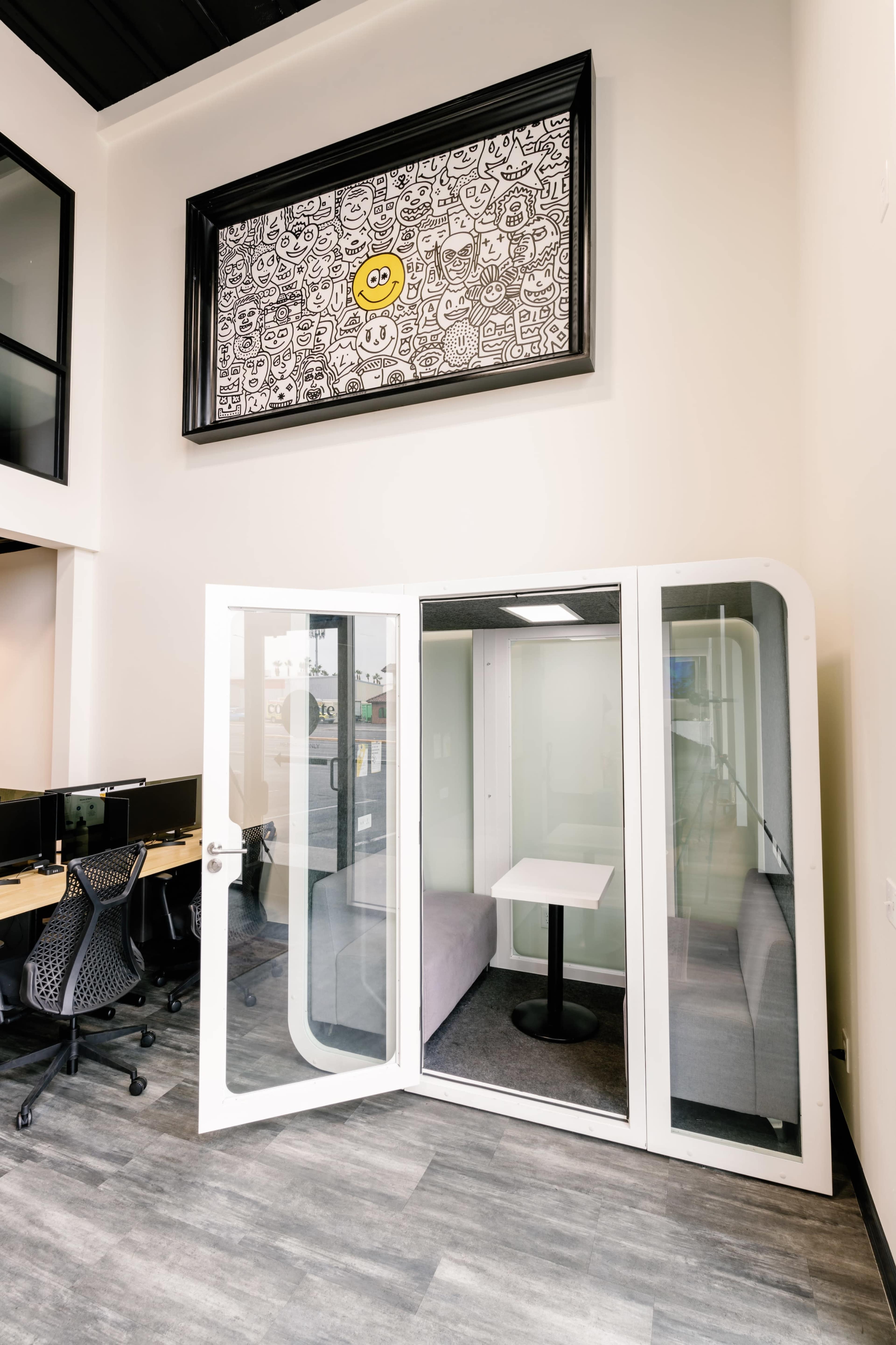 A modern glass-walled meeting pod with a table inside, situated in an office space featuring computer desks and a large framed artwork above.