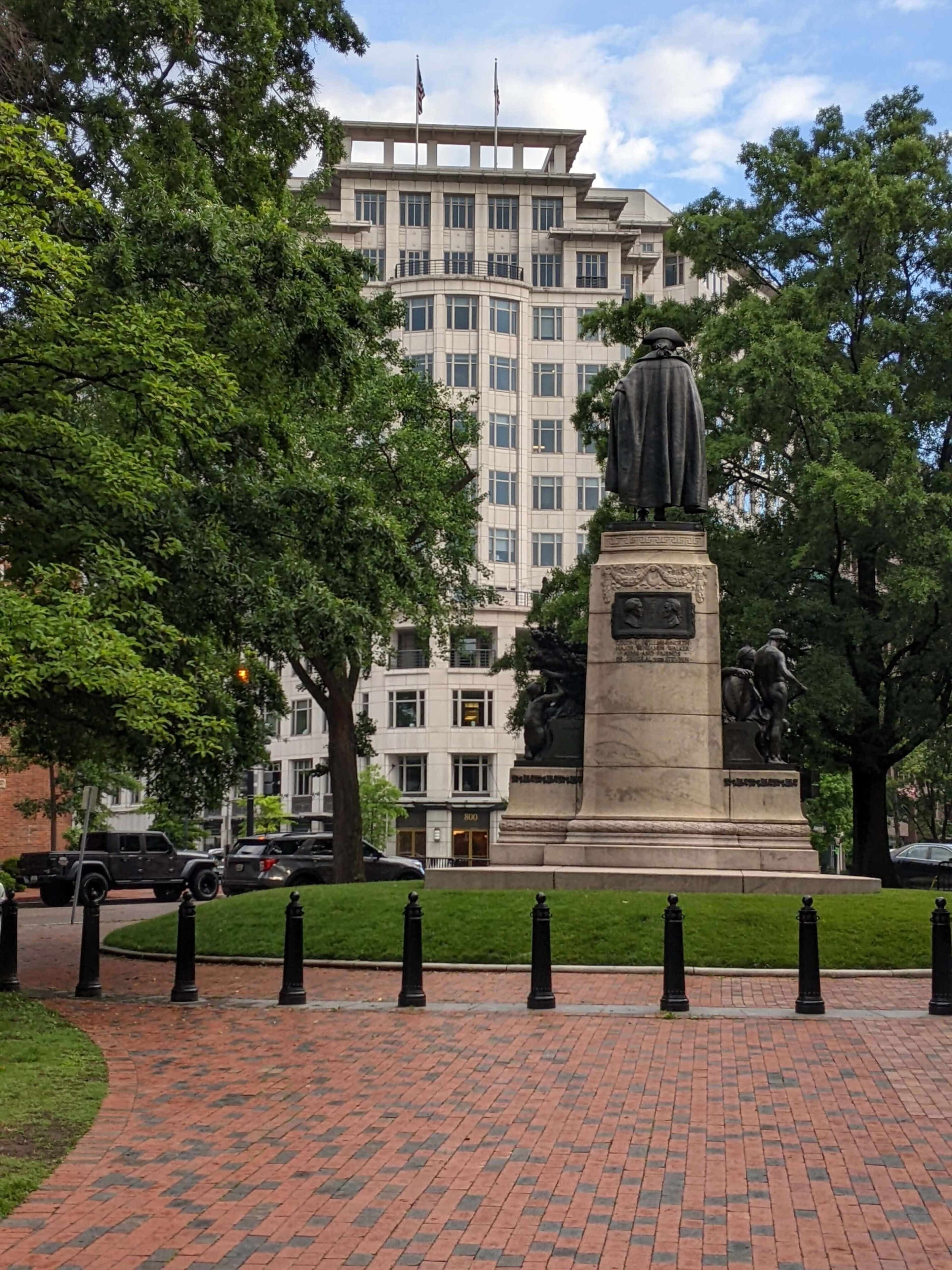 The image shows a statue on a stone pedestal surrounded by greenery and brick pathways, with a modern building in the background.