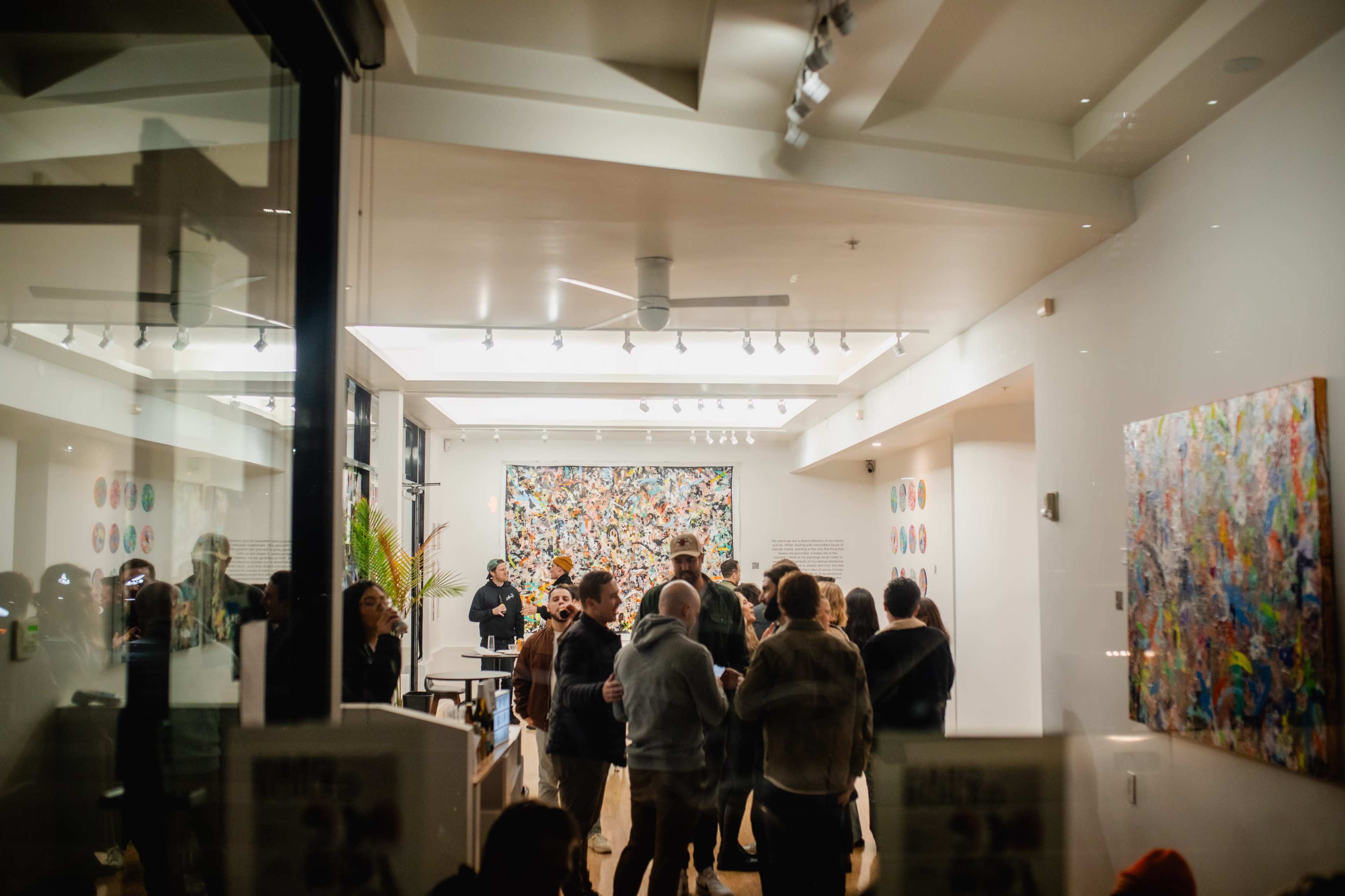 A group of people gathers in a brightly lit art gallery featuring colorful abstract artwork on the walls.