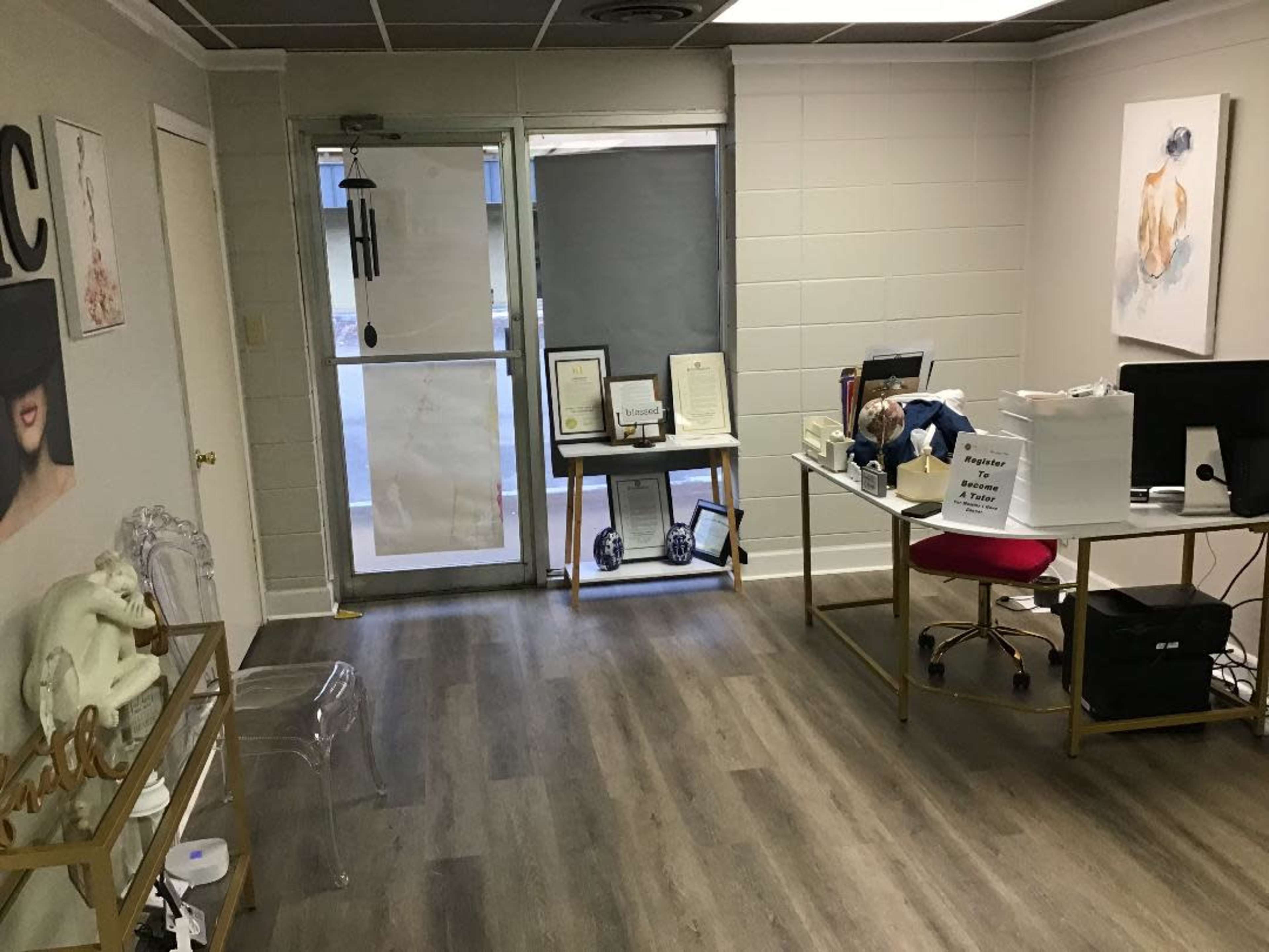 A small office space with a reception area, including a desk with a computer, framed documents on a stand, and natural light coming through a door with privacy film.