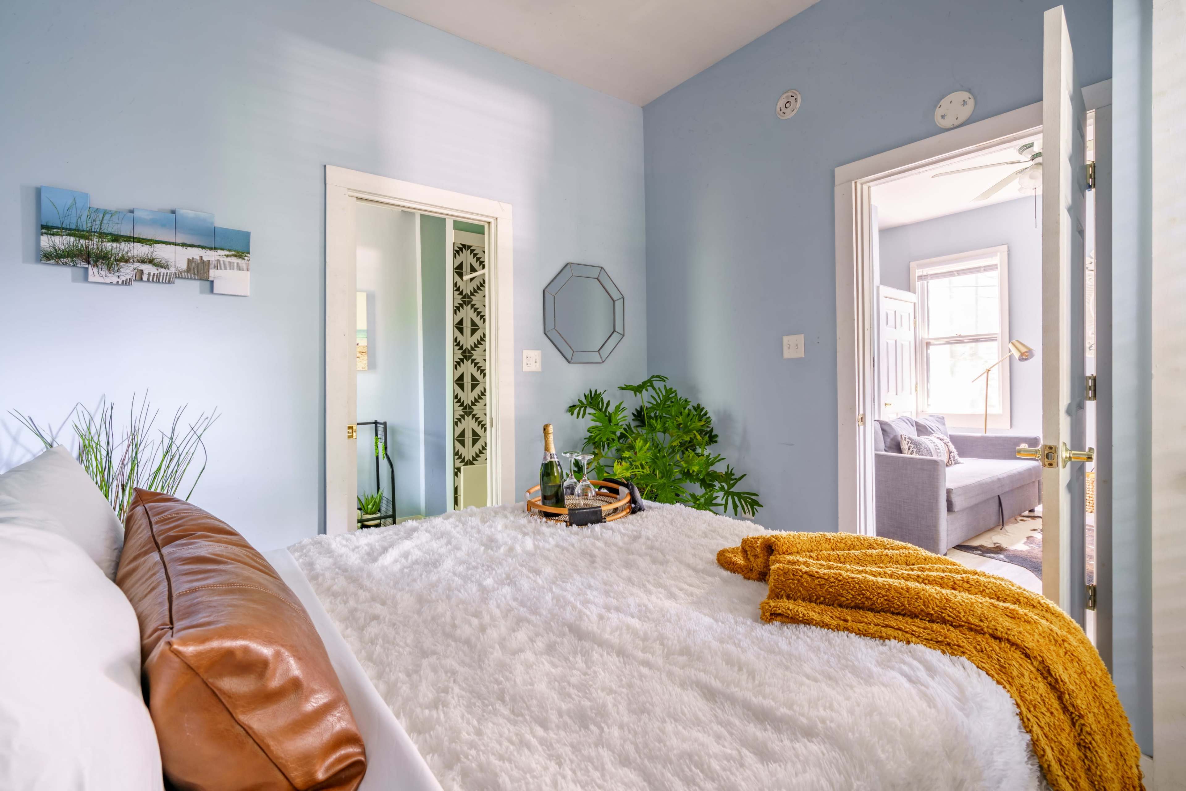 The image shows a cozy bedroom with a fluffy white bedspread, a brown leather pillow, and a small tray with bottles on the bed, alongside a view of a plant and an open door leading to another room.