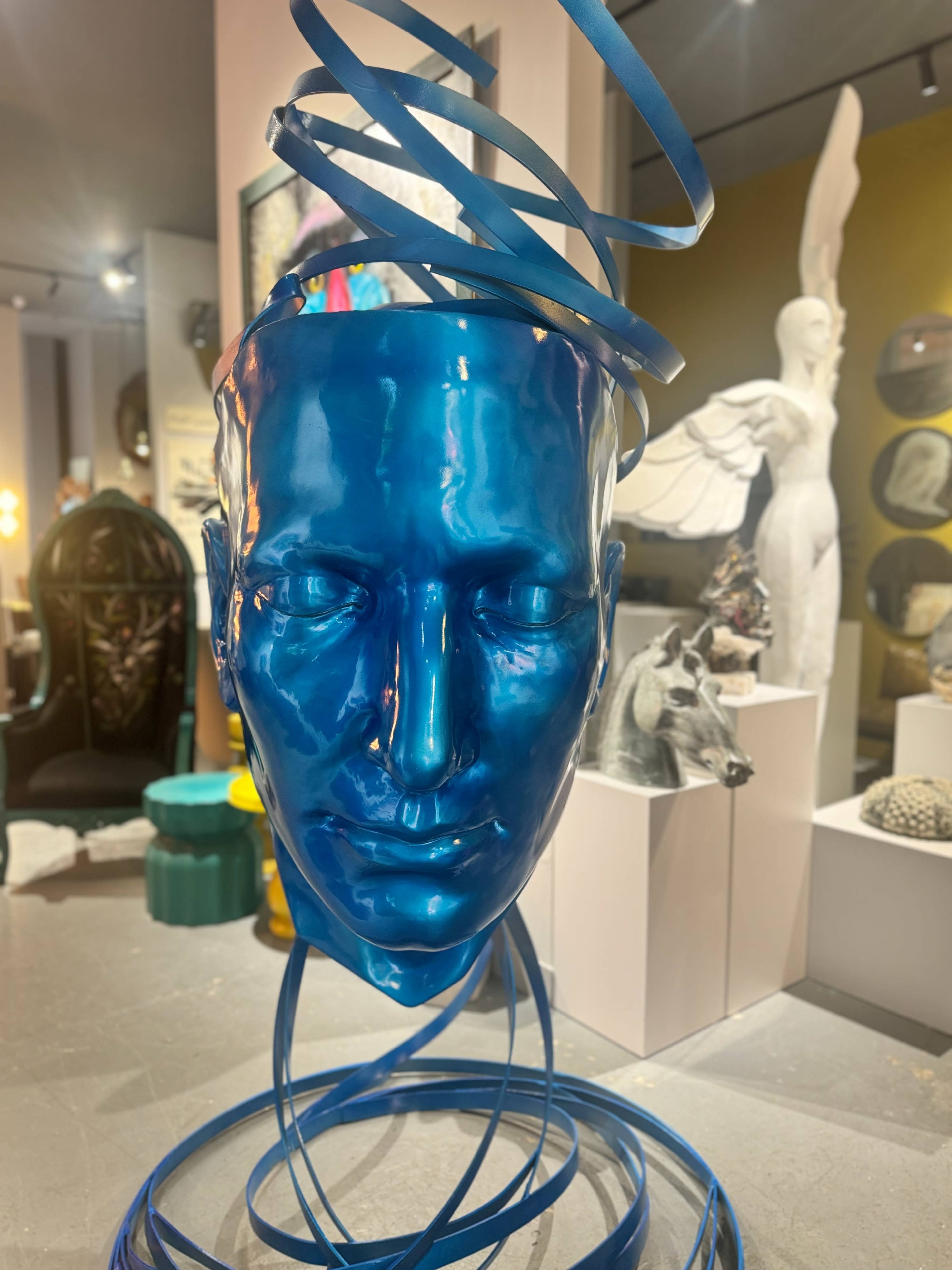 The image features a large blue head sculpture with spiraling blue ribbons around it, set in a gallery that displays various art pieces.