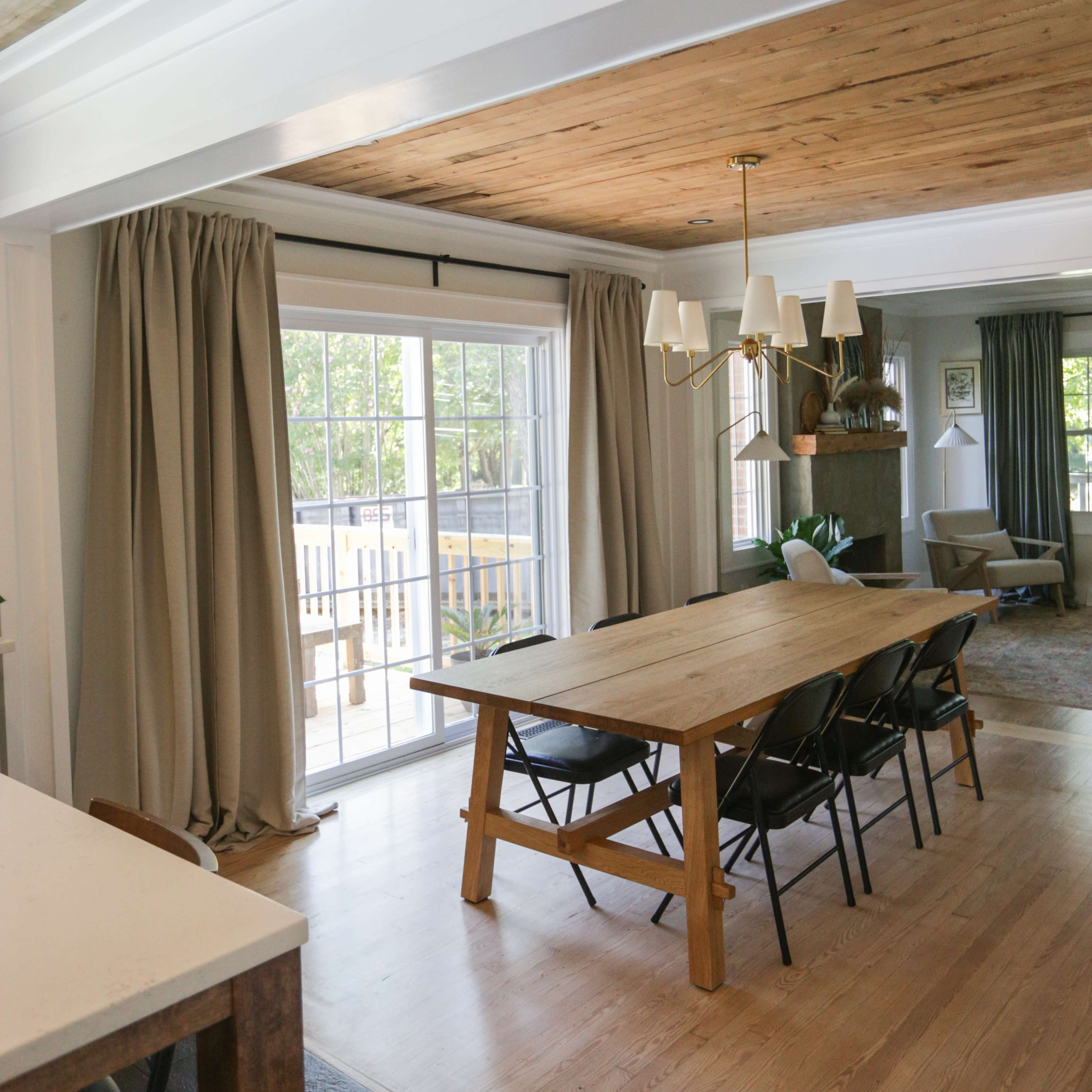 A spacious dining area features a long wooden table surrounded by black chairs, with large windows draped in curtains allowing natural light to flood the room.