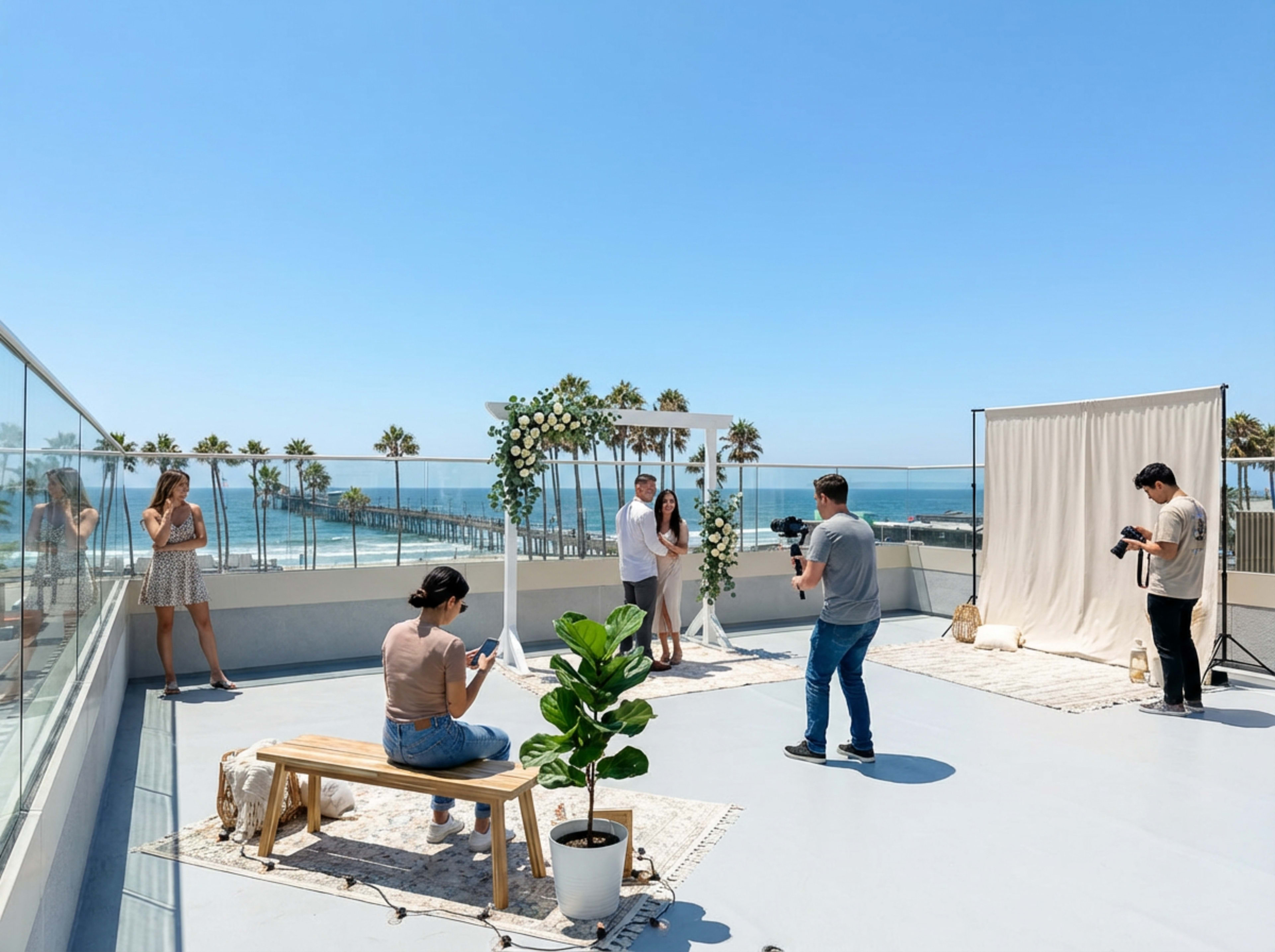 A group of people participates in a photo shoot on a sunny rooftop with palm trees and the ocean in the background.