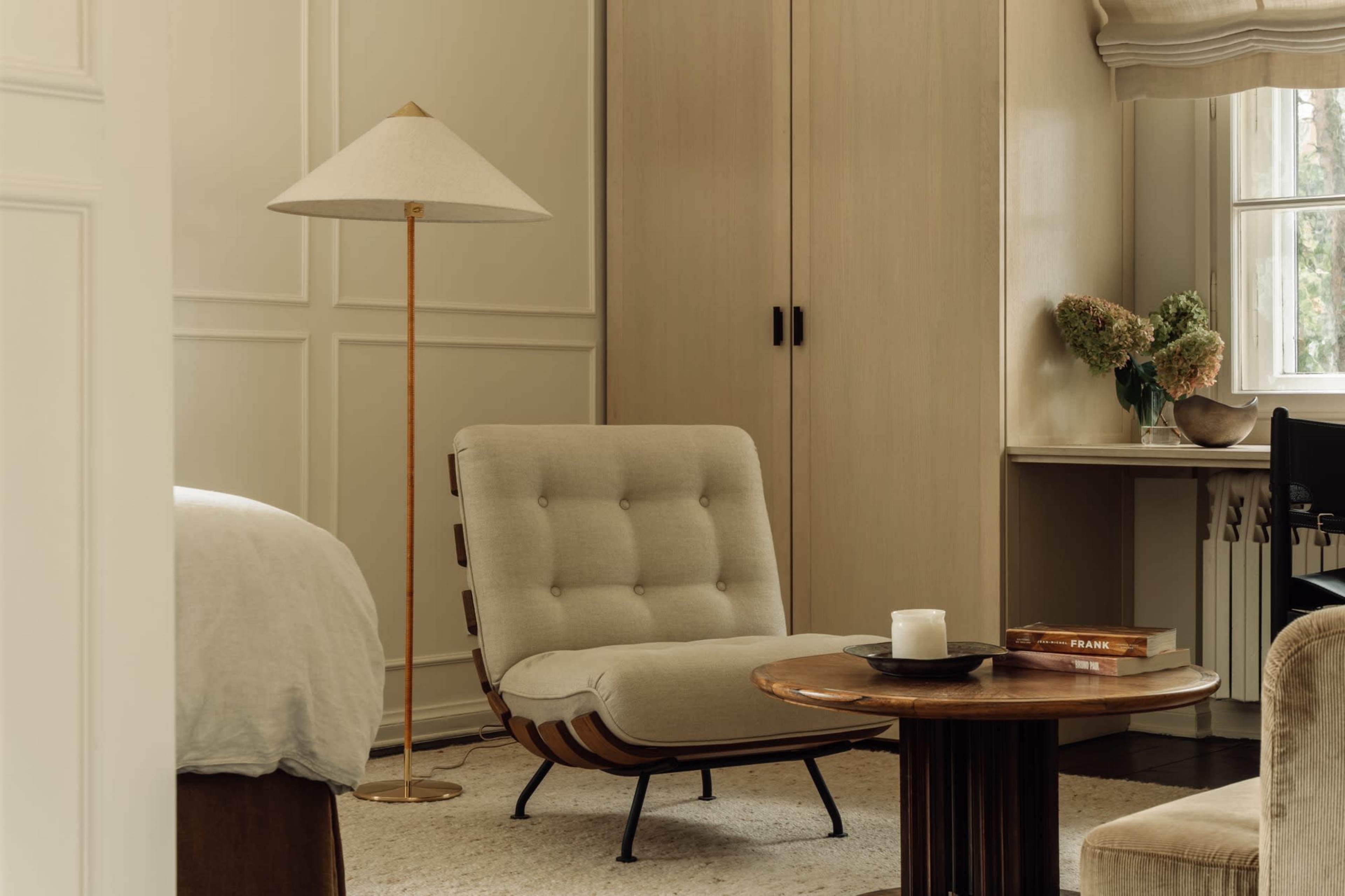 A cozy corner of a room featuring a beige lounge chair, a wooden side table with a candle, a floor lamp, and a vase with flowers on a desk against a light-colored wall.