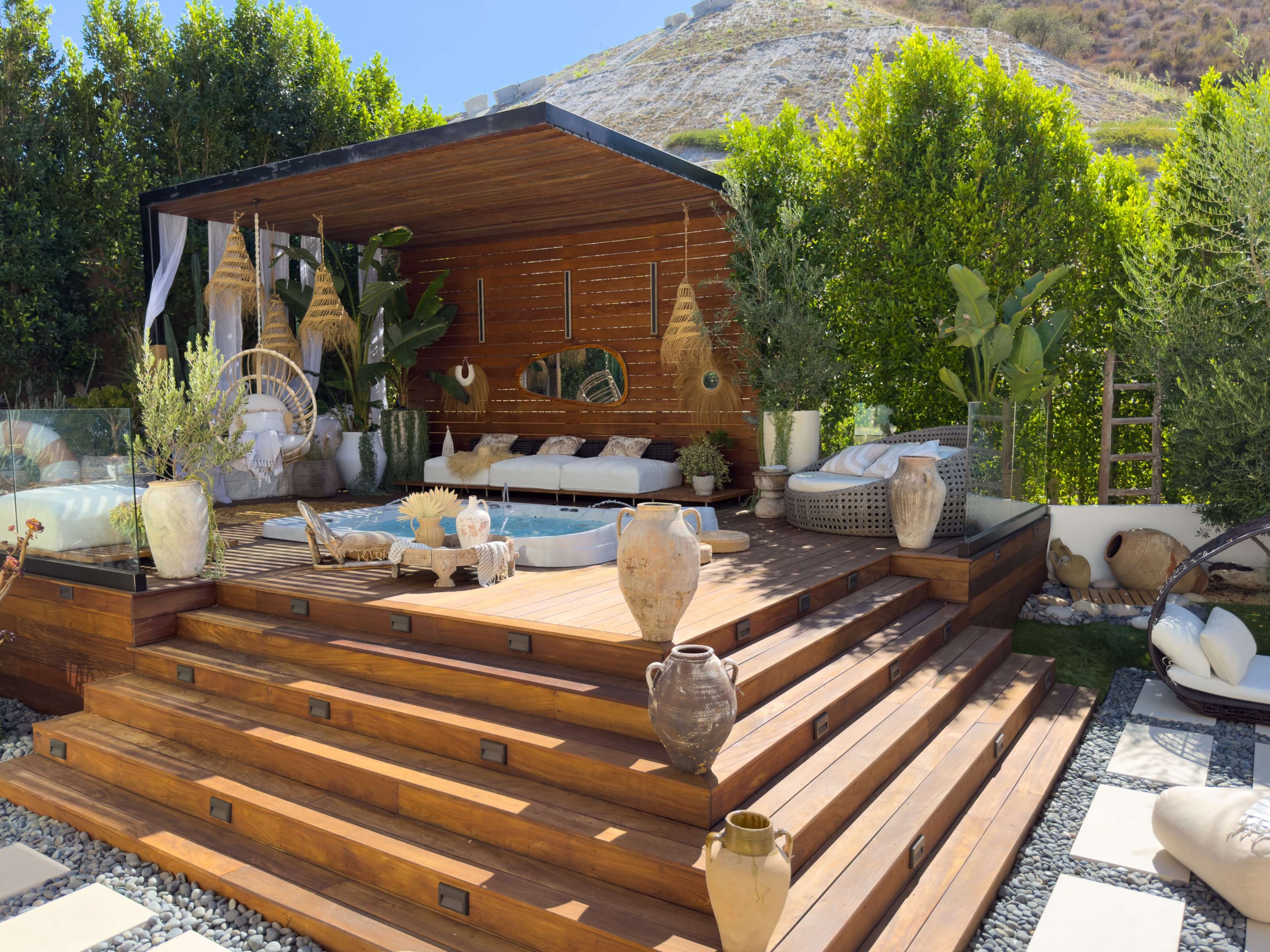 The image features a stylish outdoor lounge area with wooden decking, comfortable seating, decorative vases, and lush green plants surrounded by a serene landscape.