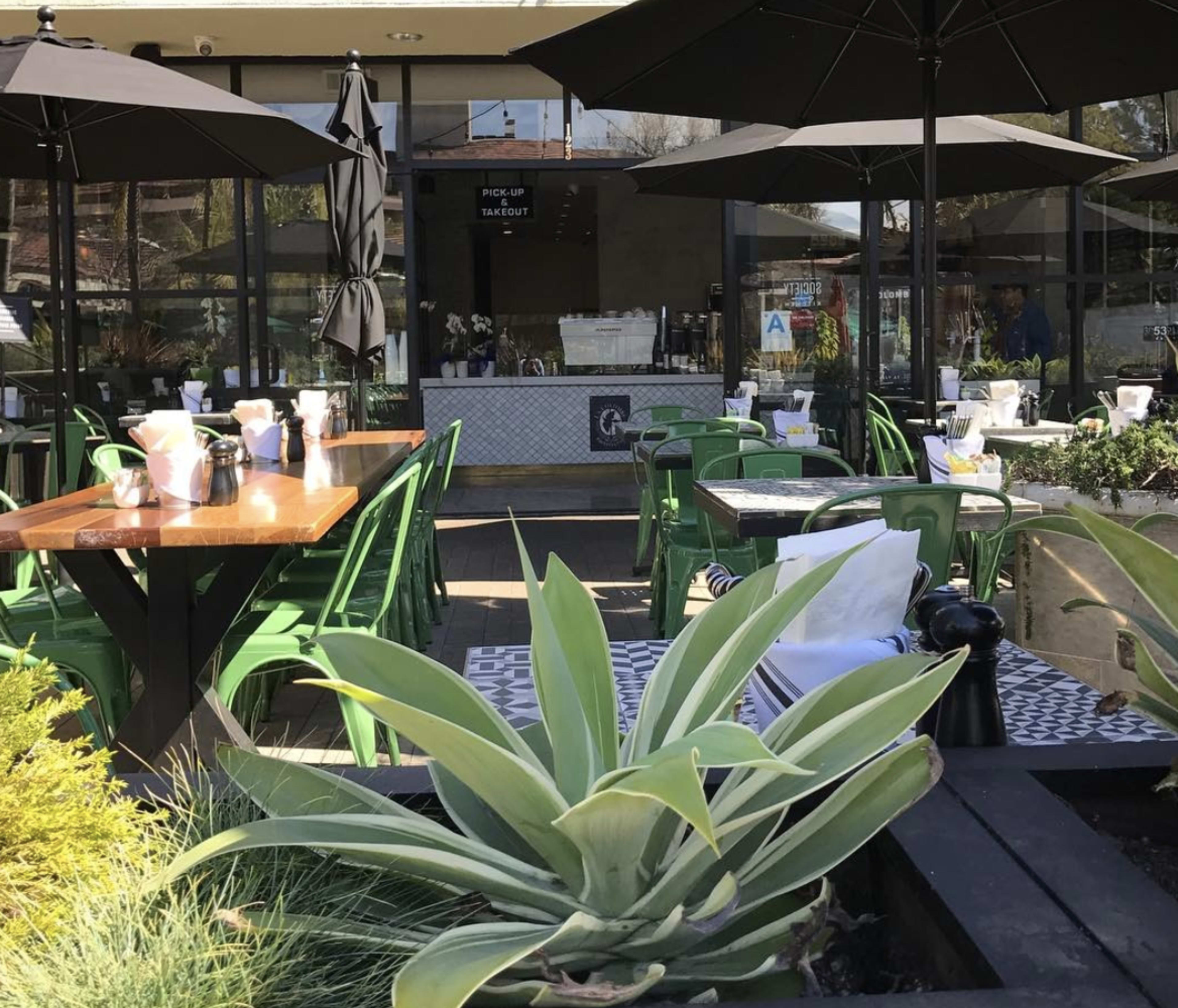 A modern outdoor dining area features green chairs and tables shaded by black umbrellas, surrounded by potted plants and decorative tile.
