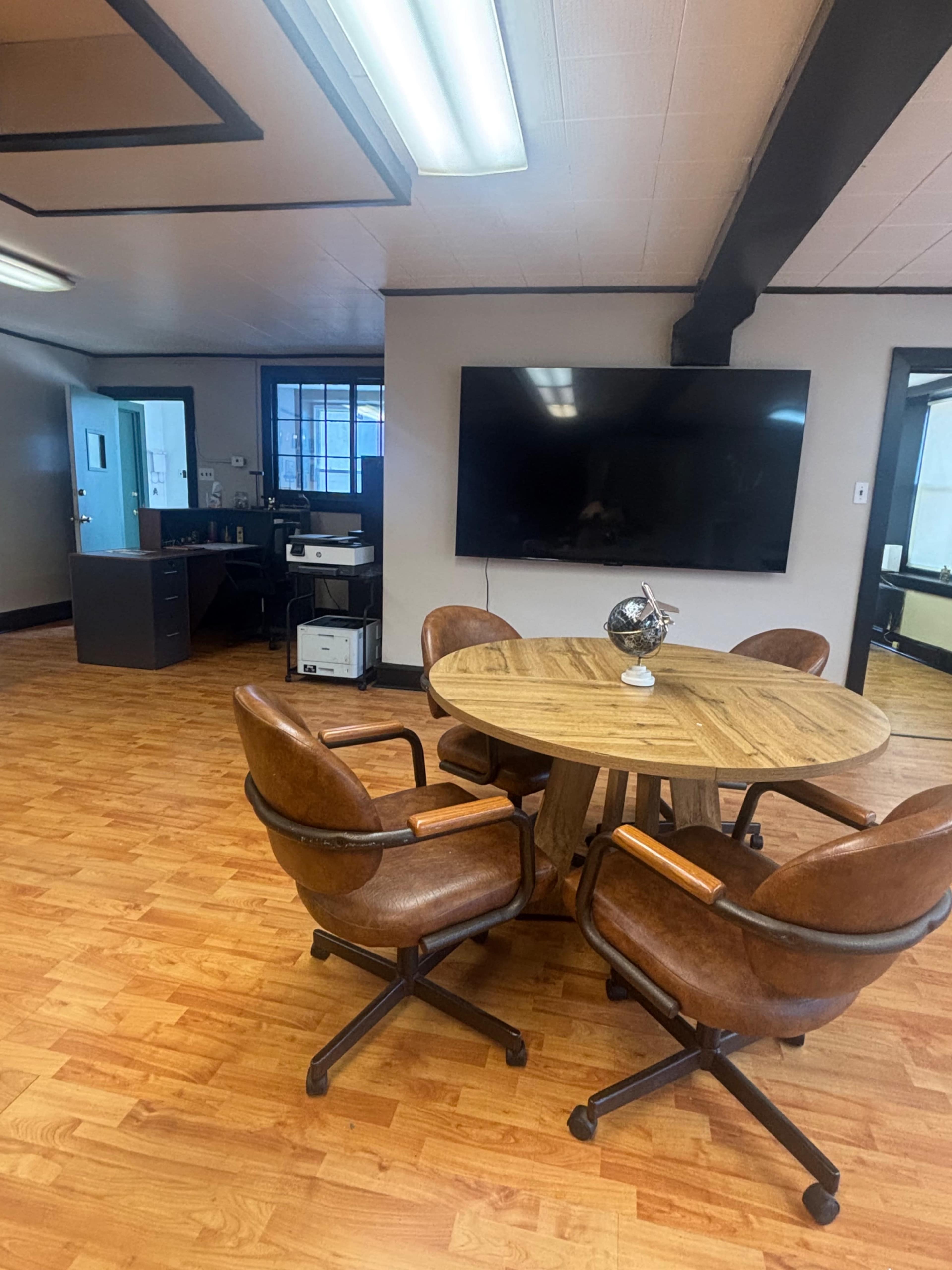 The image shows a spacious office area featuring a round wooden table surrounded by four brown leather chairs, with a large television and office equipment in the background.