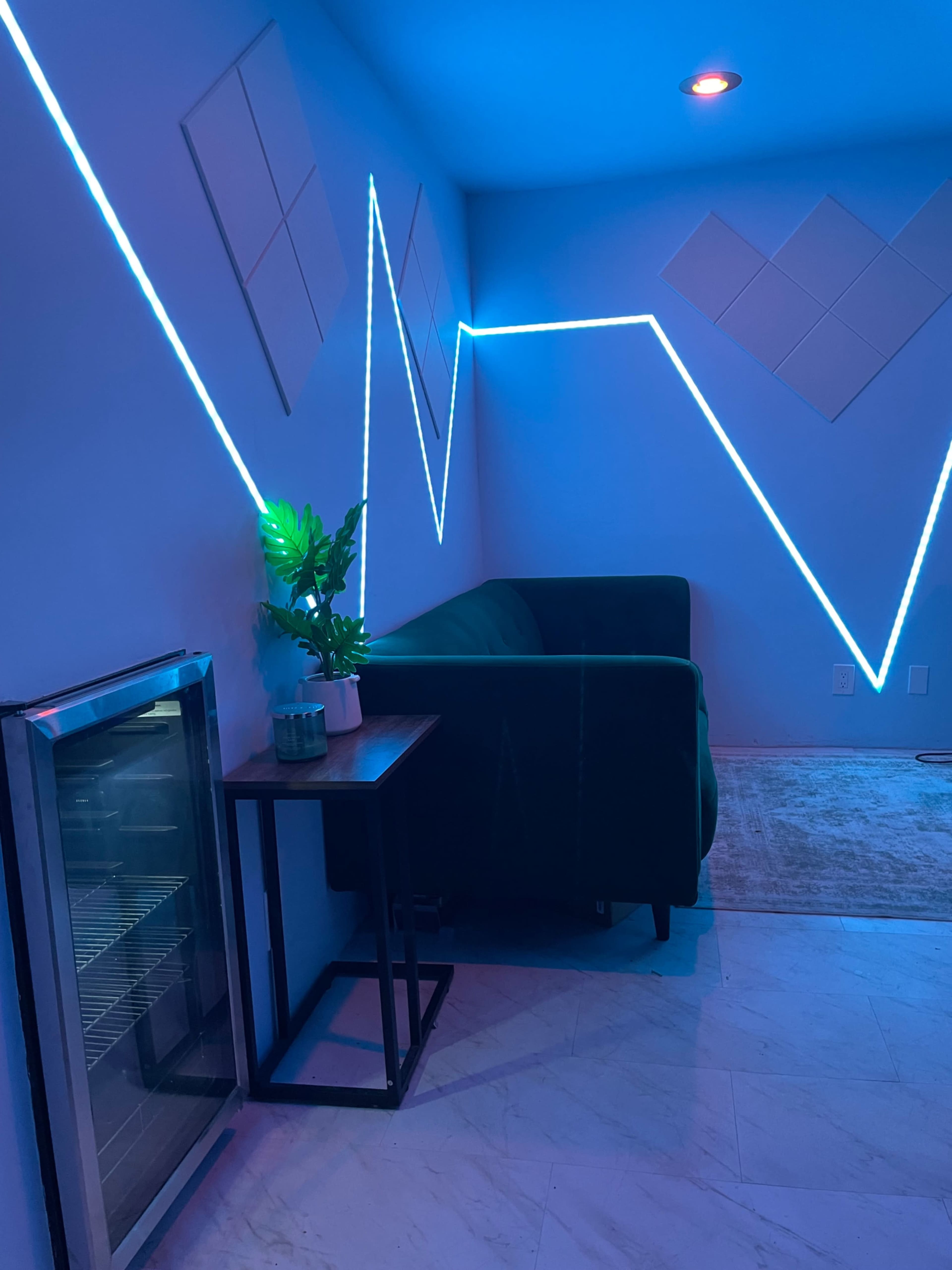 The image features a cozy corner of a room with a green couch, a small table, a plant, and blue LED lights illuminating the walls in a zigzag pattern.