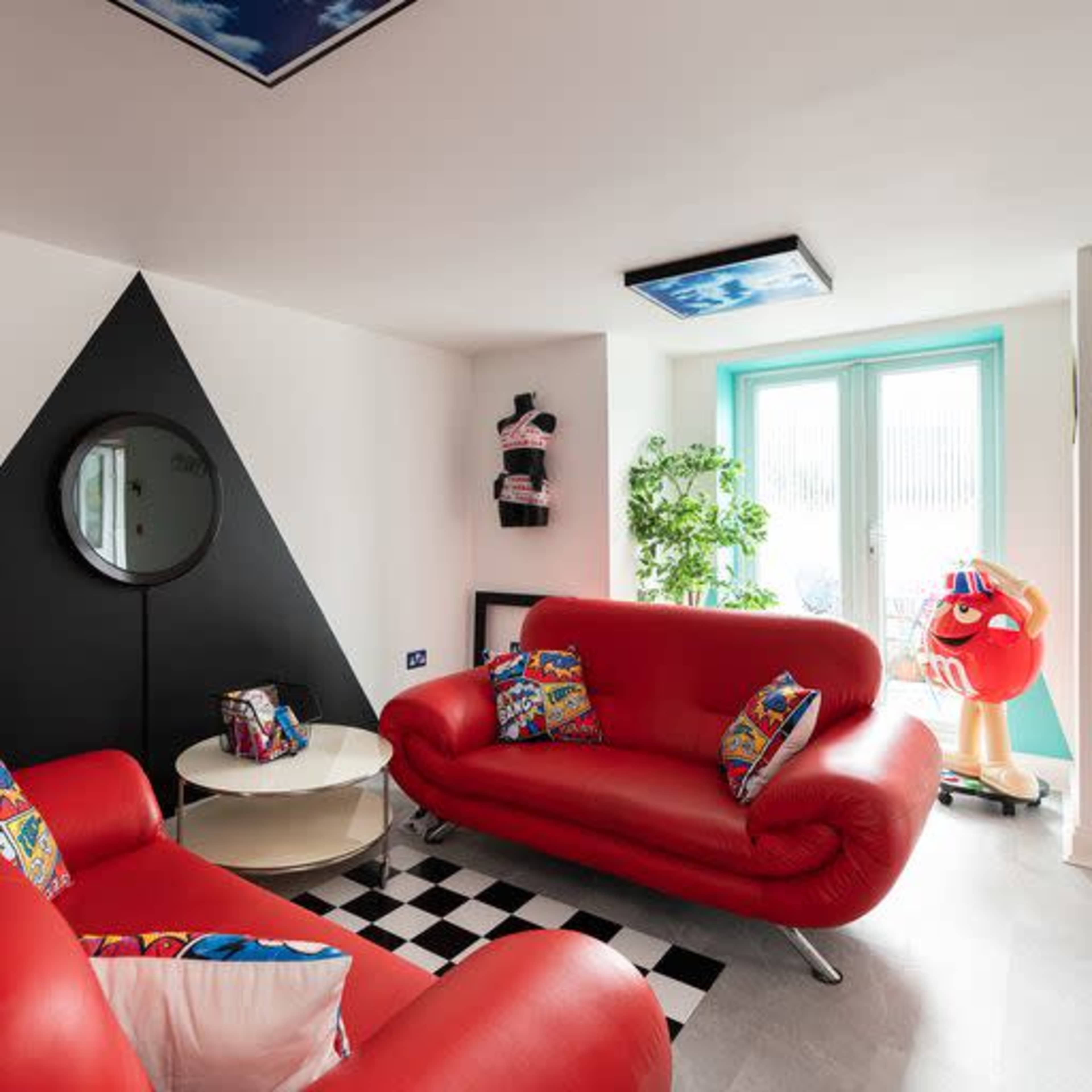 A modern living room with a red sofa, a black-and-white checkered floor, and a large wall mirror, featuring a geometric black wall design and large windows opening to a patio.