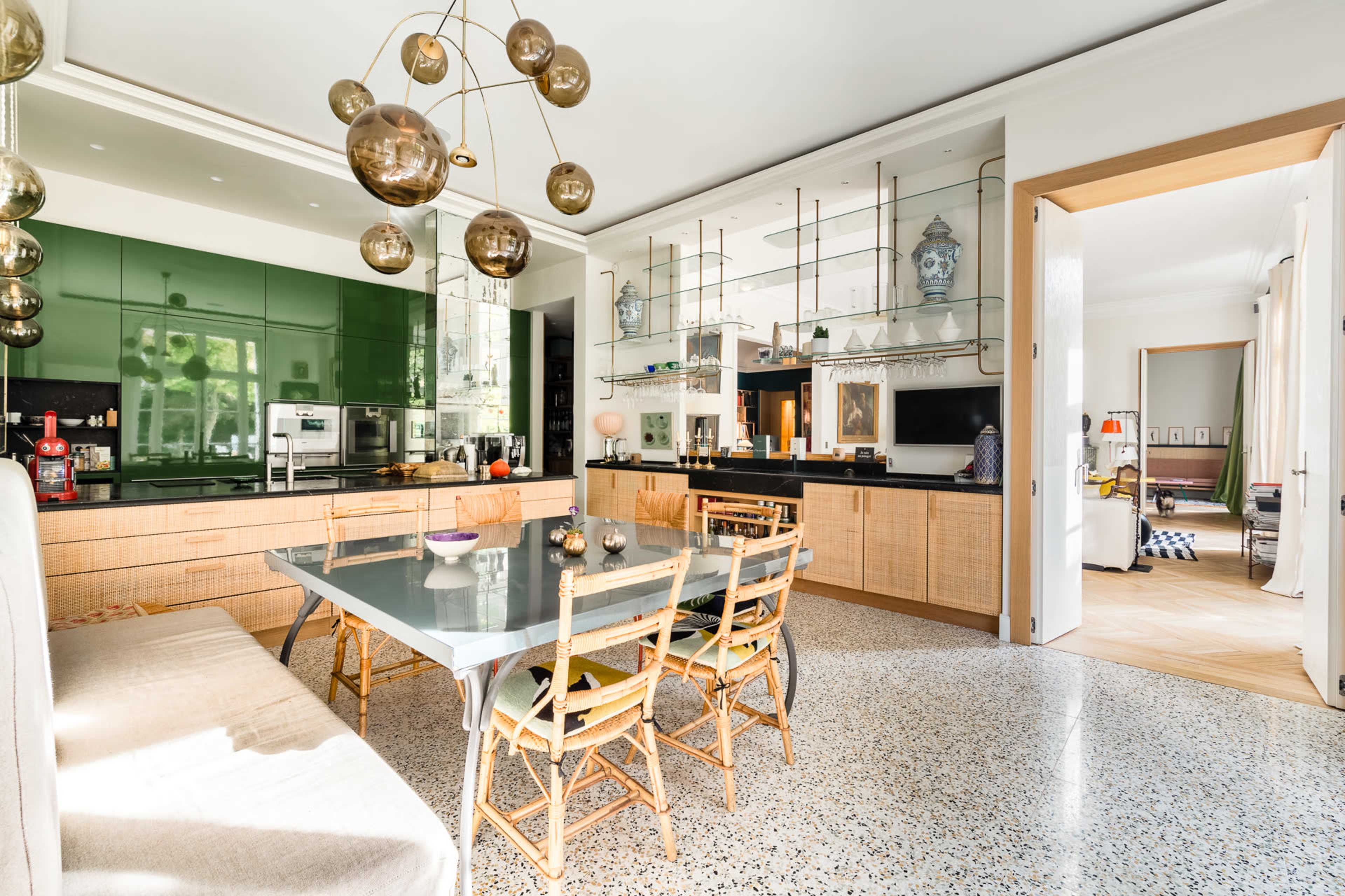 A modern dining area features a long table surrounded by wooden chairs, with a green kitchen backdrop and decorative shelves above.