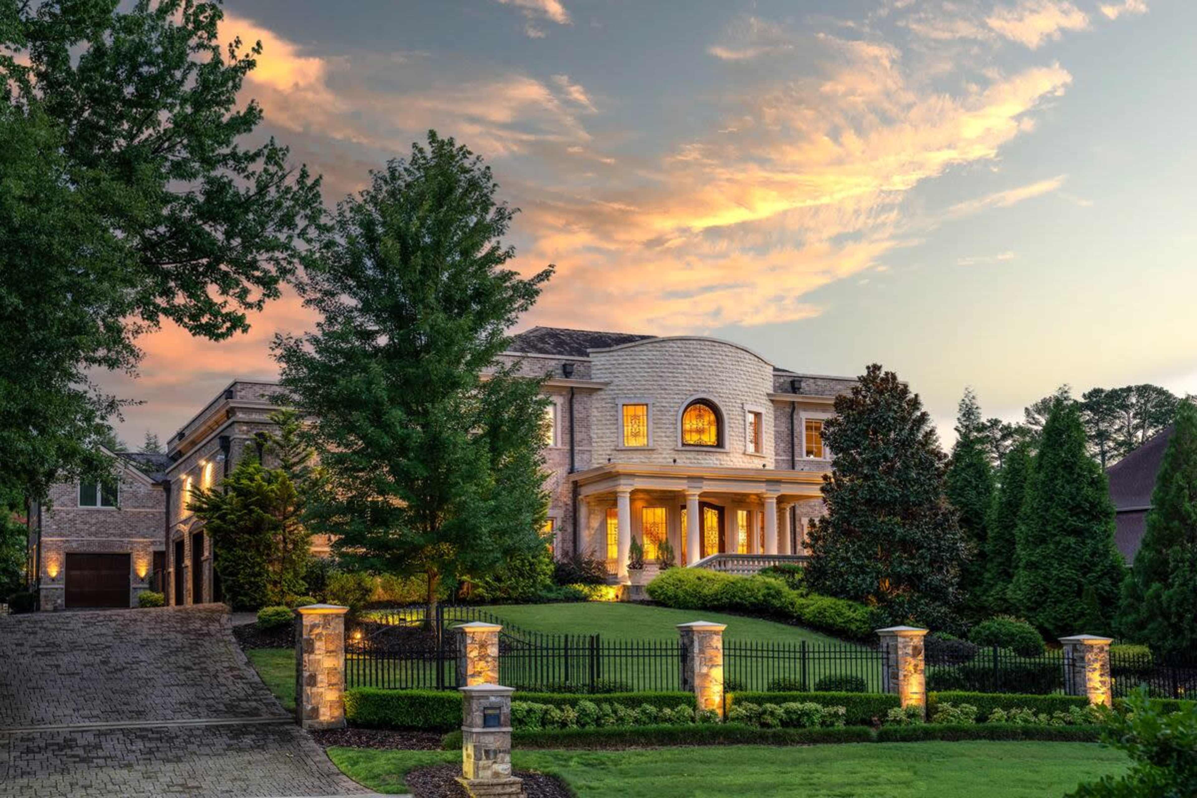 A large, elegant house with prominent architectural features is illuminated at dusk, set on a manicured lawn with a stone driveway and decorative fencing.