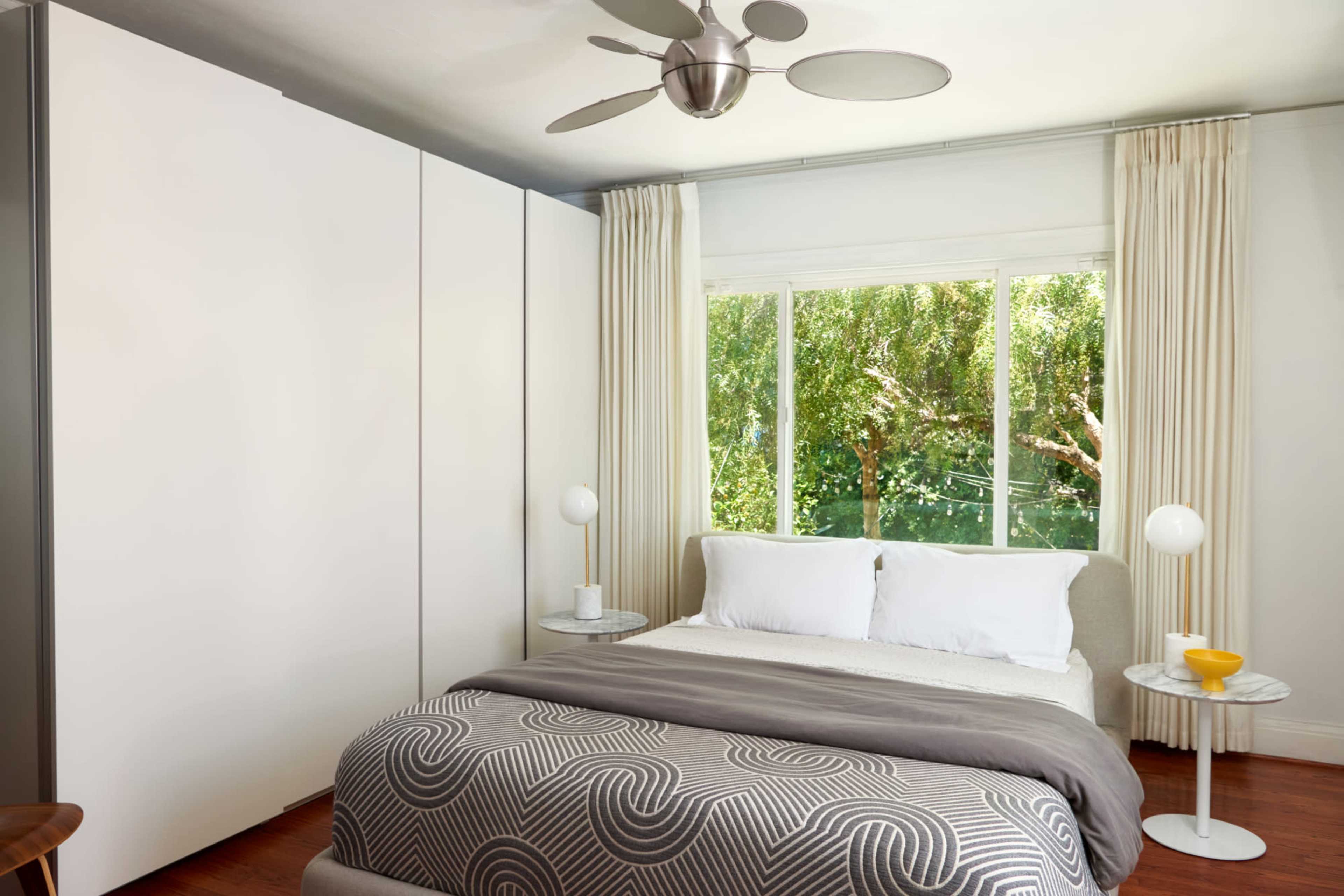 The bedroom features a bed with white linens, a patterned gray blanket, and two side tables with lamps, set against a backdrop of large windows overlooking greenery.