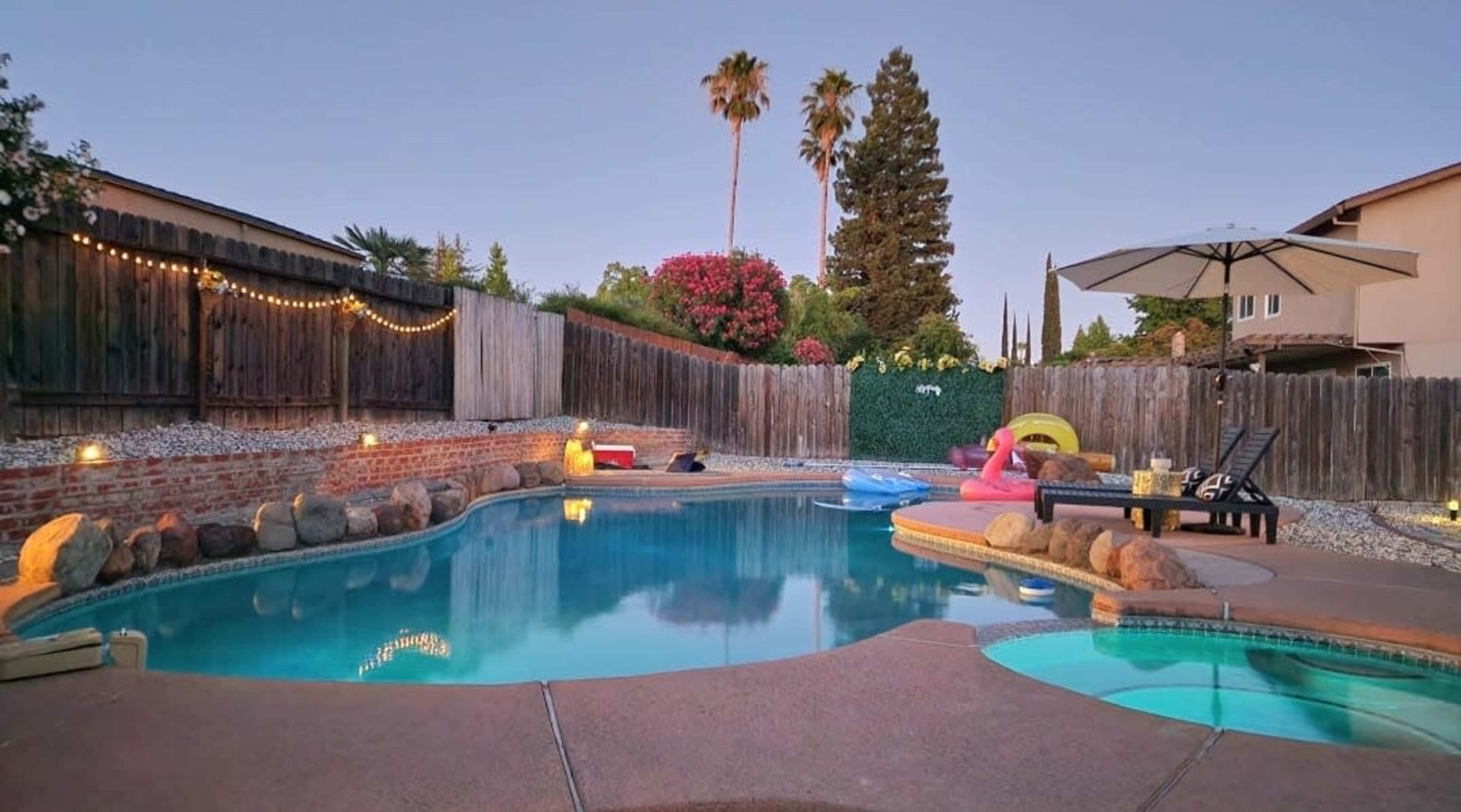 The image shows a backyard pool surrounded by trees and decorative lights, with lounge chairs and floating toys on the water.
