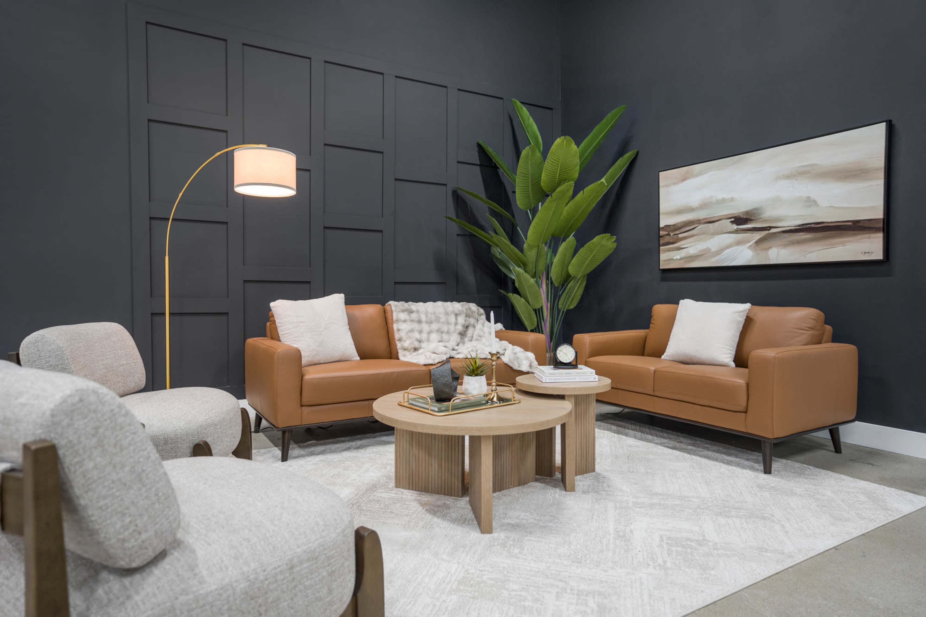 The image shows a modern living room setup featuring two brown leather sofas, a round wooden coffee table, a grey rug, and a large potted plant against a dark wall.