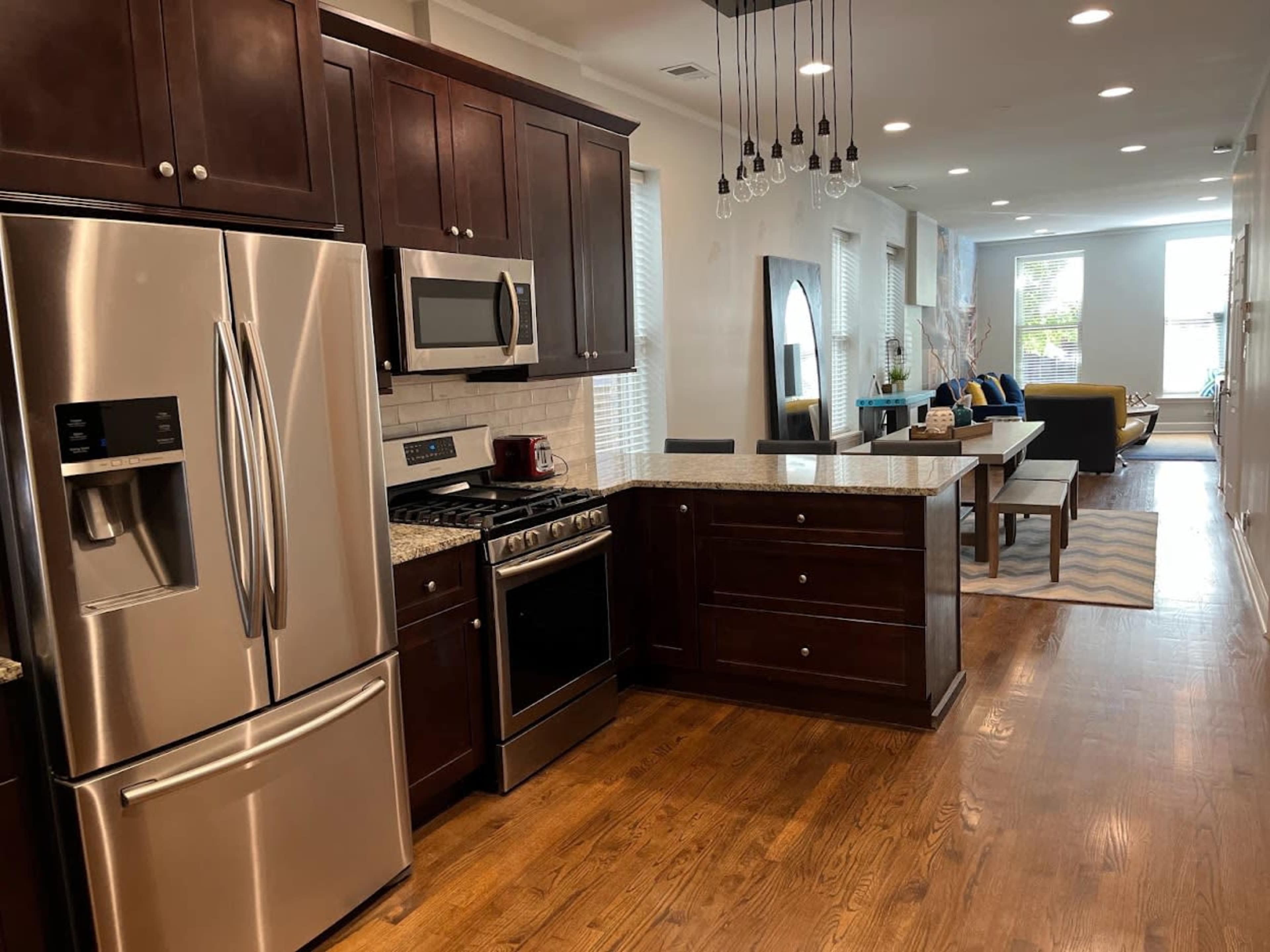 A modern kitchen with dark wooden cabinets, stainless steel appliances, and an island countertop, adjacent to a living area featuring a sofa and a coffee table.