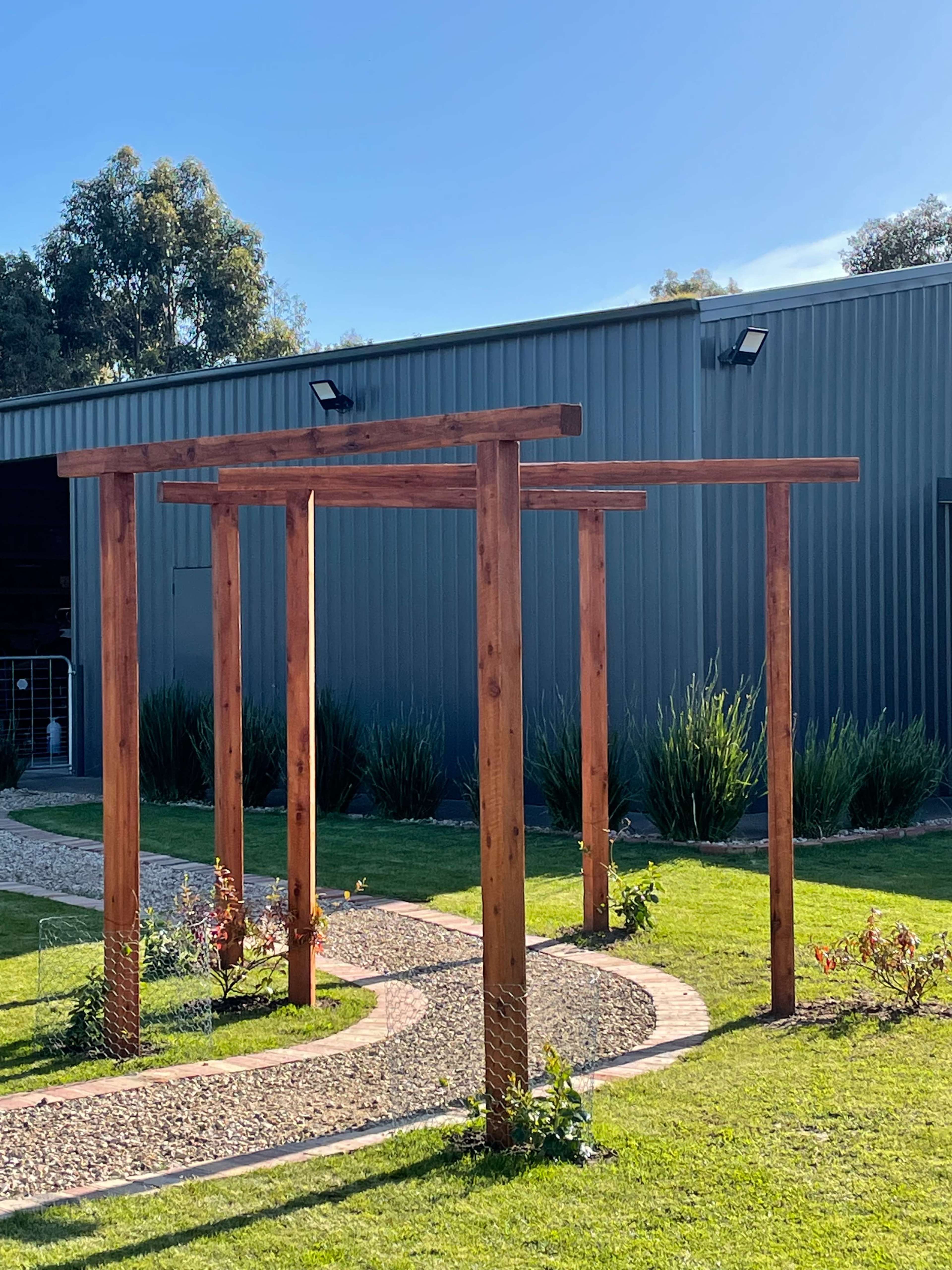 A wooden pergola structure with four vertical posts, set on a lawn beside a gravel path and a grey metal building.