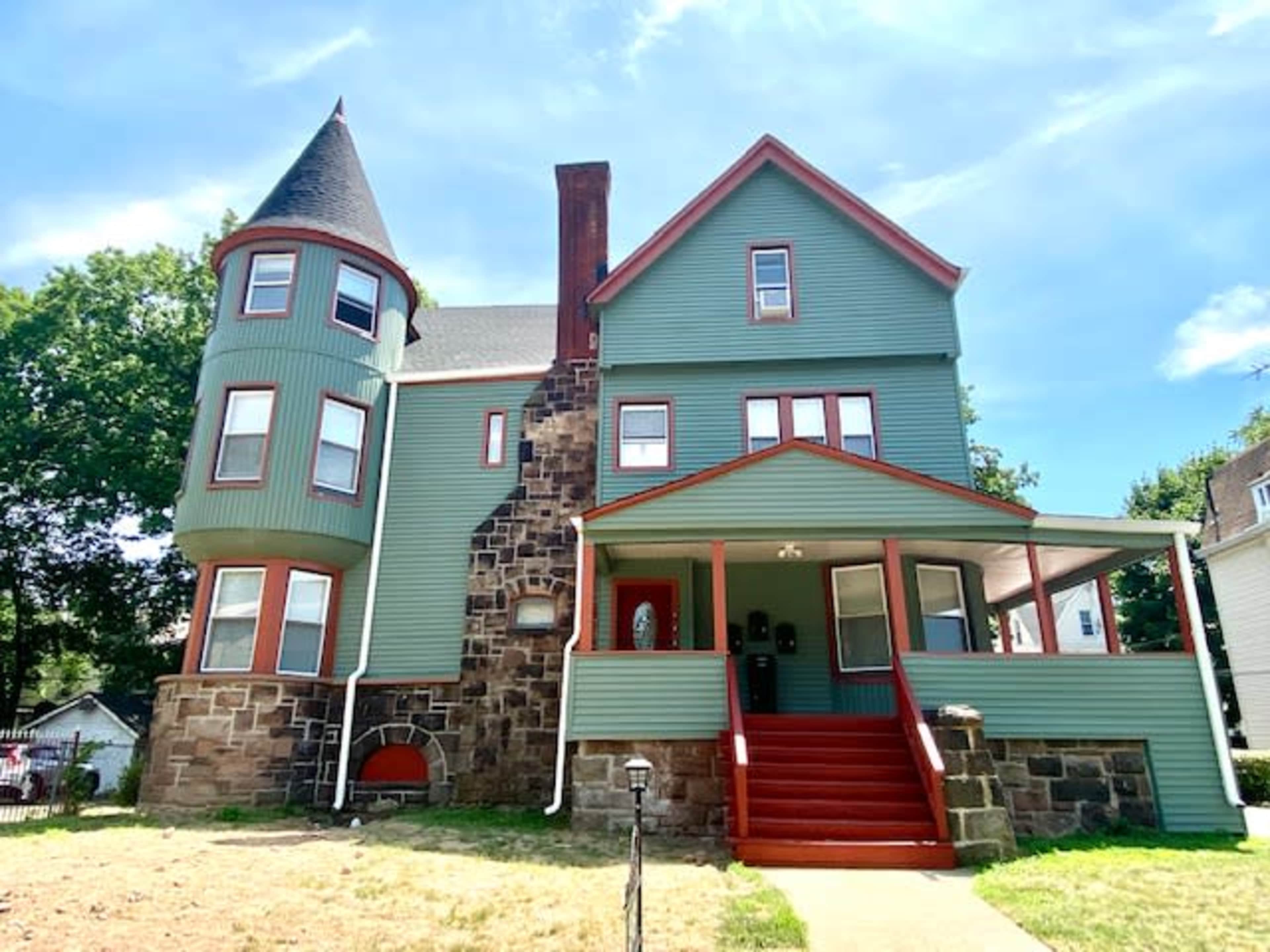 The image shows a multi-story house with a stone foundation, green siding, and a distinctive turret.