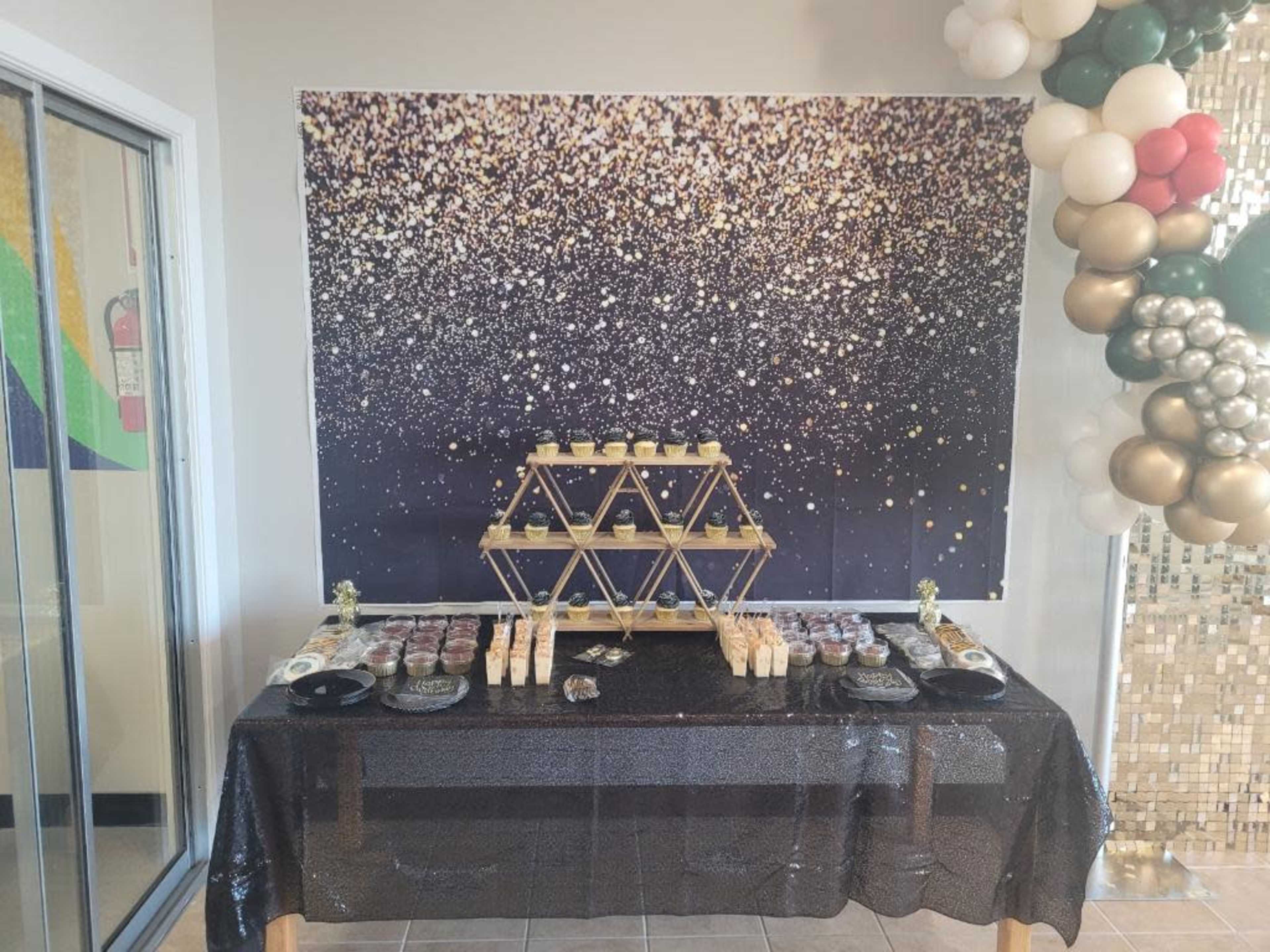 The image shows a decorated table covered with a black tablecloth, displaying an assortment of desserts, set against a sparkly gold and black backdrop.