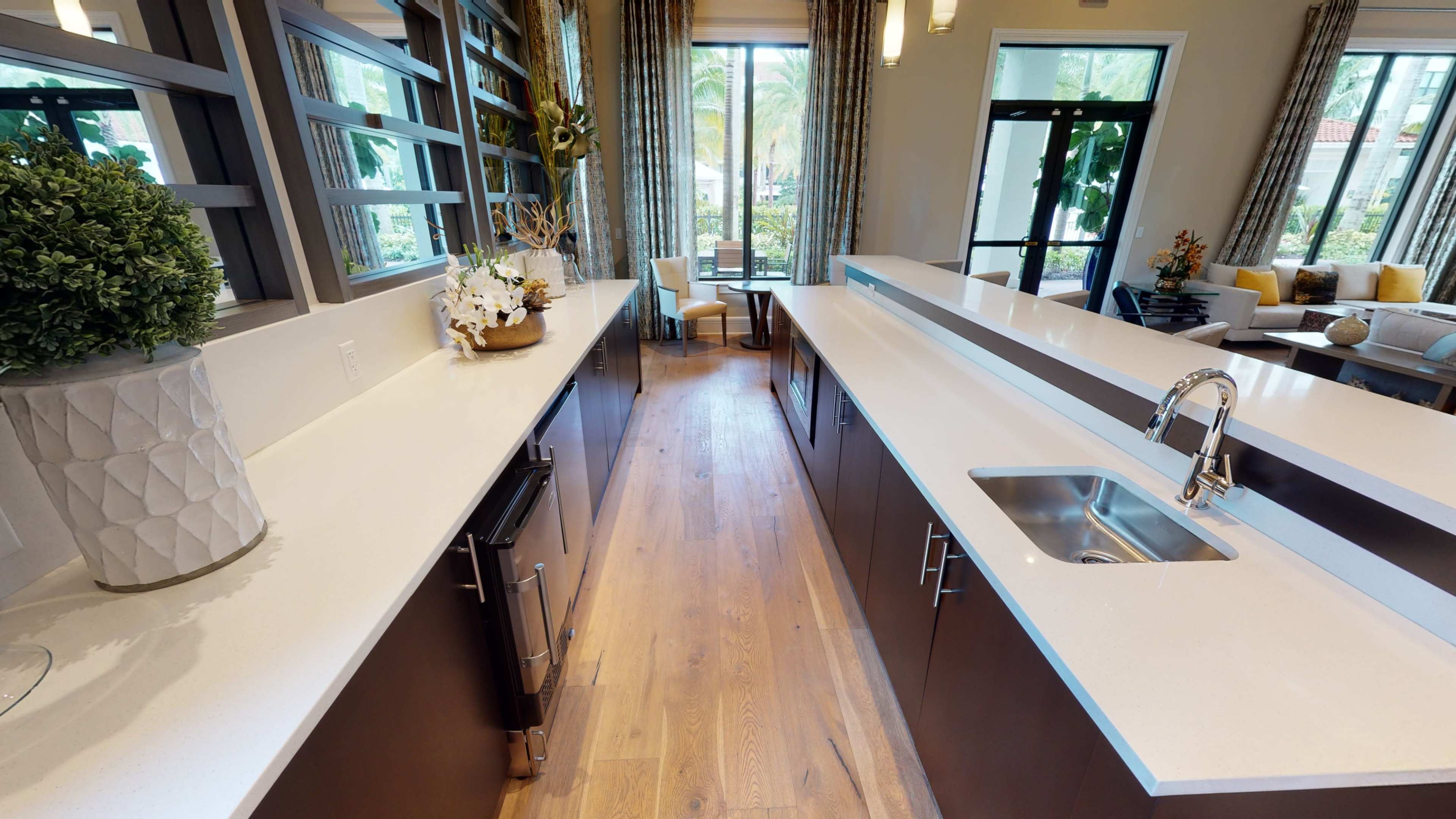 The image shows a modern kitchen with two parallel countertops, a sink on the right, and a view of a seating area and windows in the background.