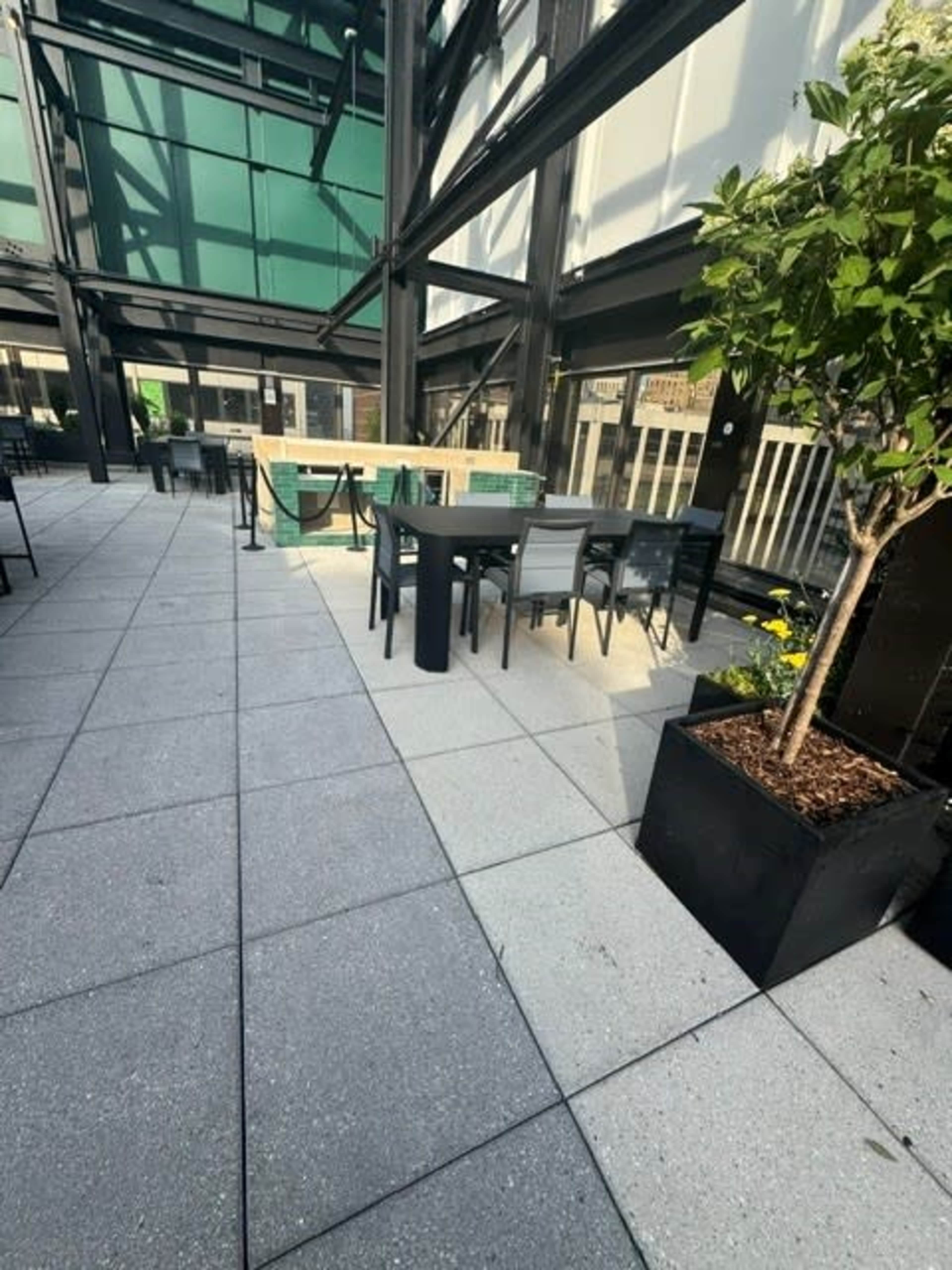 The image shows a modern outdoor dining area with tables, chairs, and potted plants under a glass roof structure.