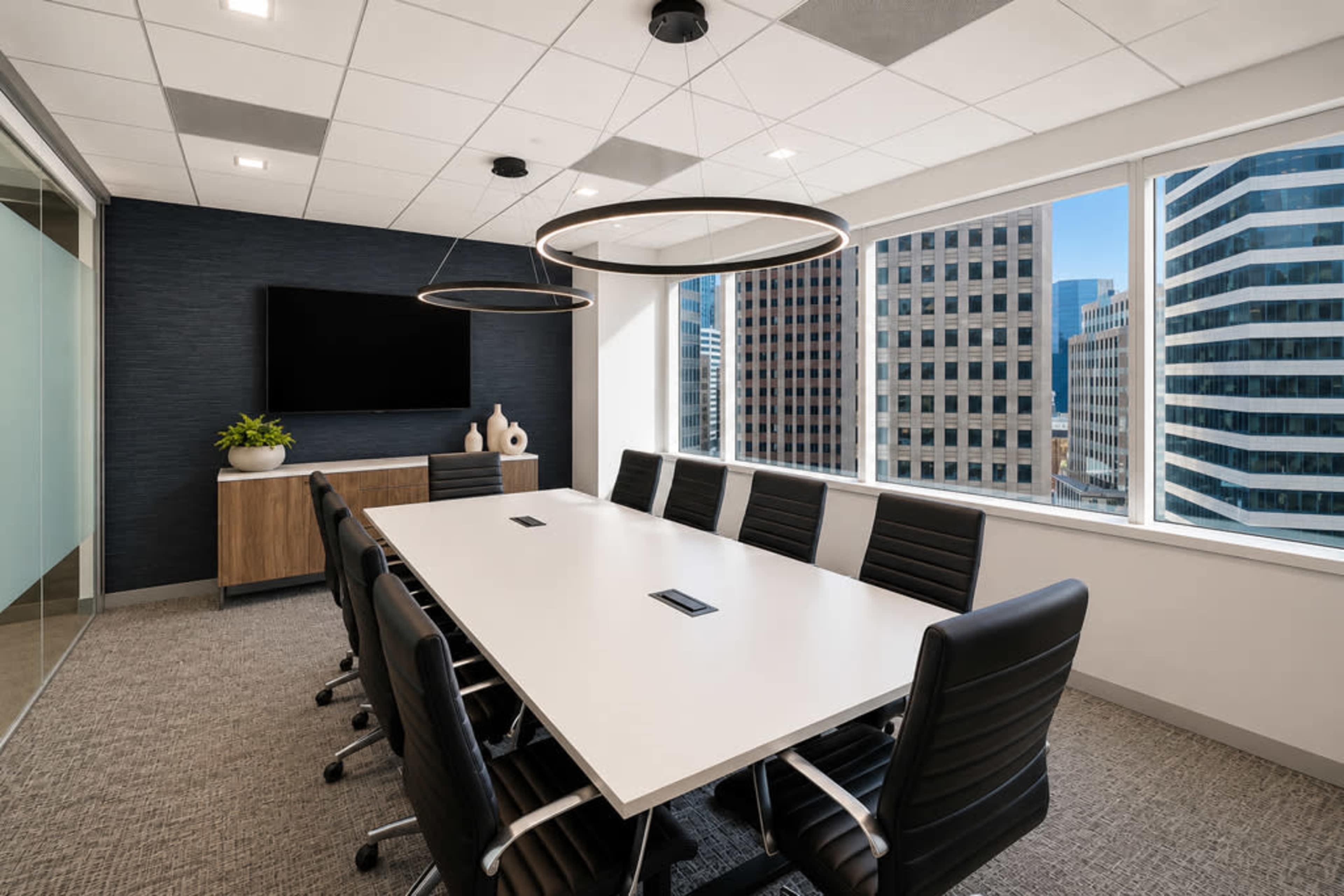 A modern conference room features a long white table surrounded by black chairs, with large windows showcasing a city skyline outside.