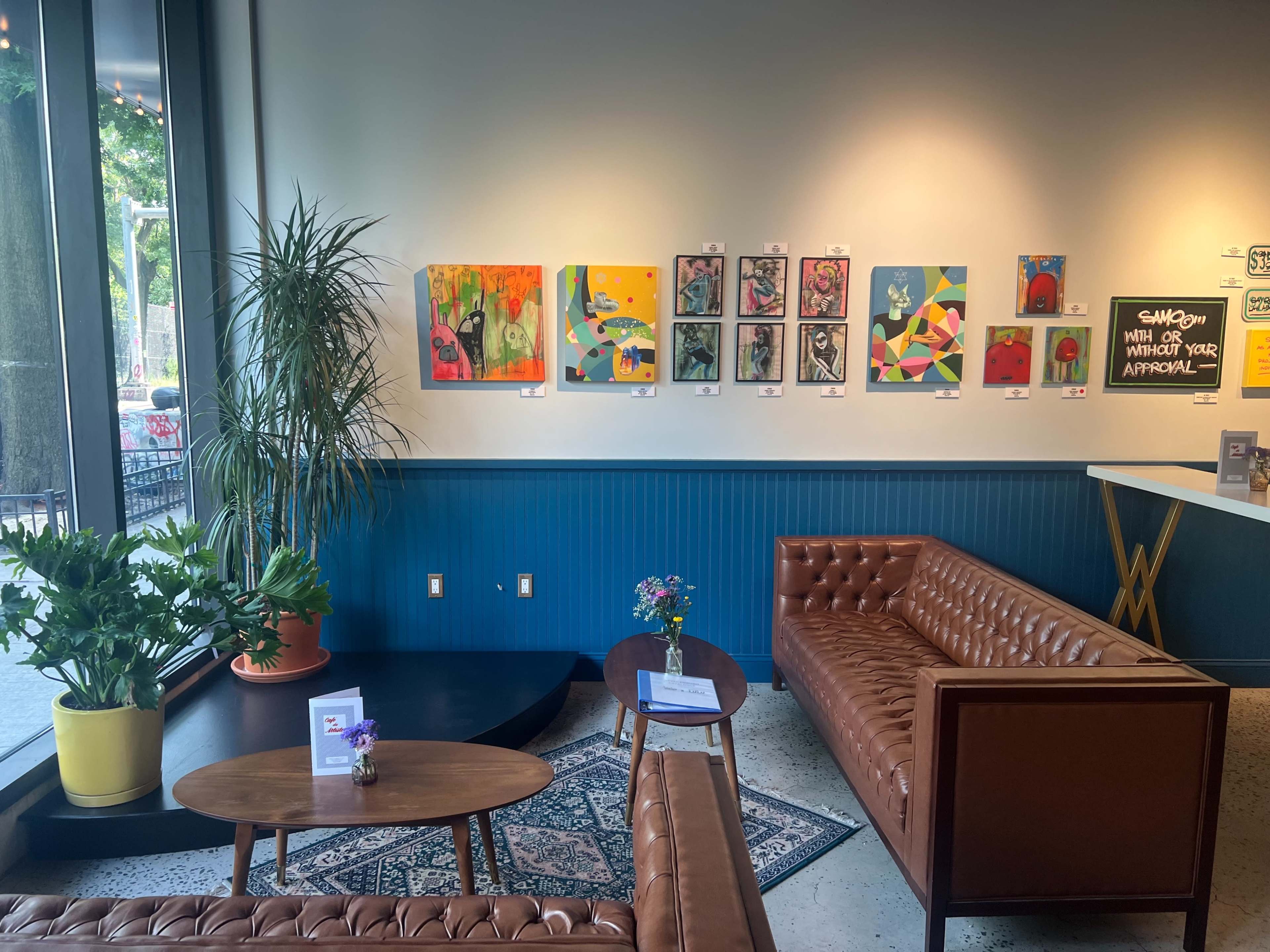 The image shows a cozy indoor space with two brown leather sofas, a small table, potted plants, and colorful artwork displayed on the walls.