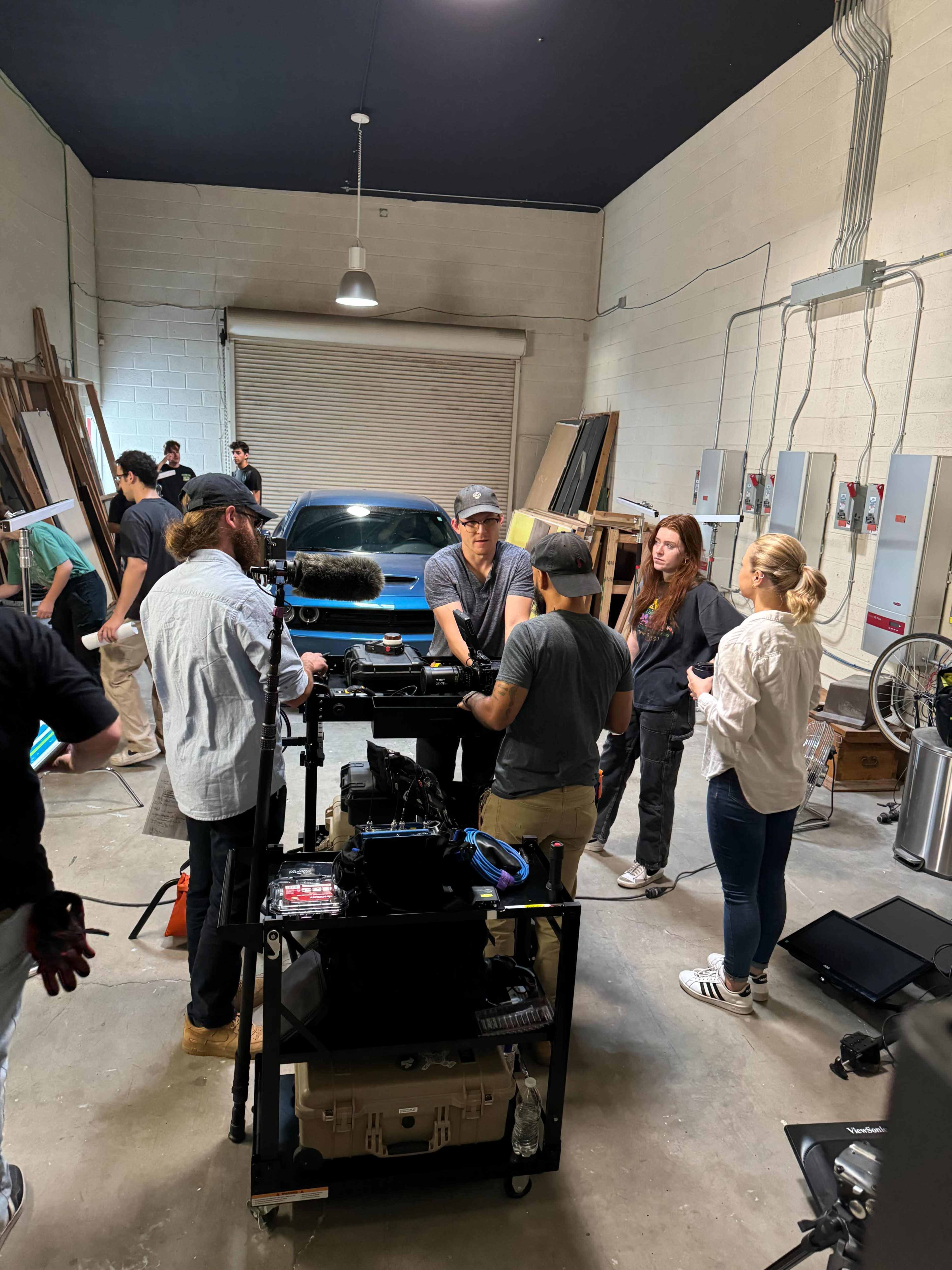 A group of people collaborates around a camera setup in a garage-like space with a car and various equipment in the background.
