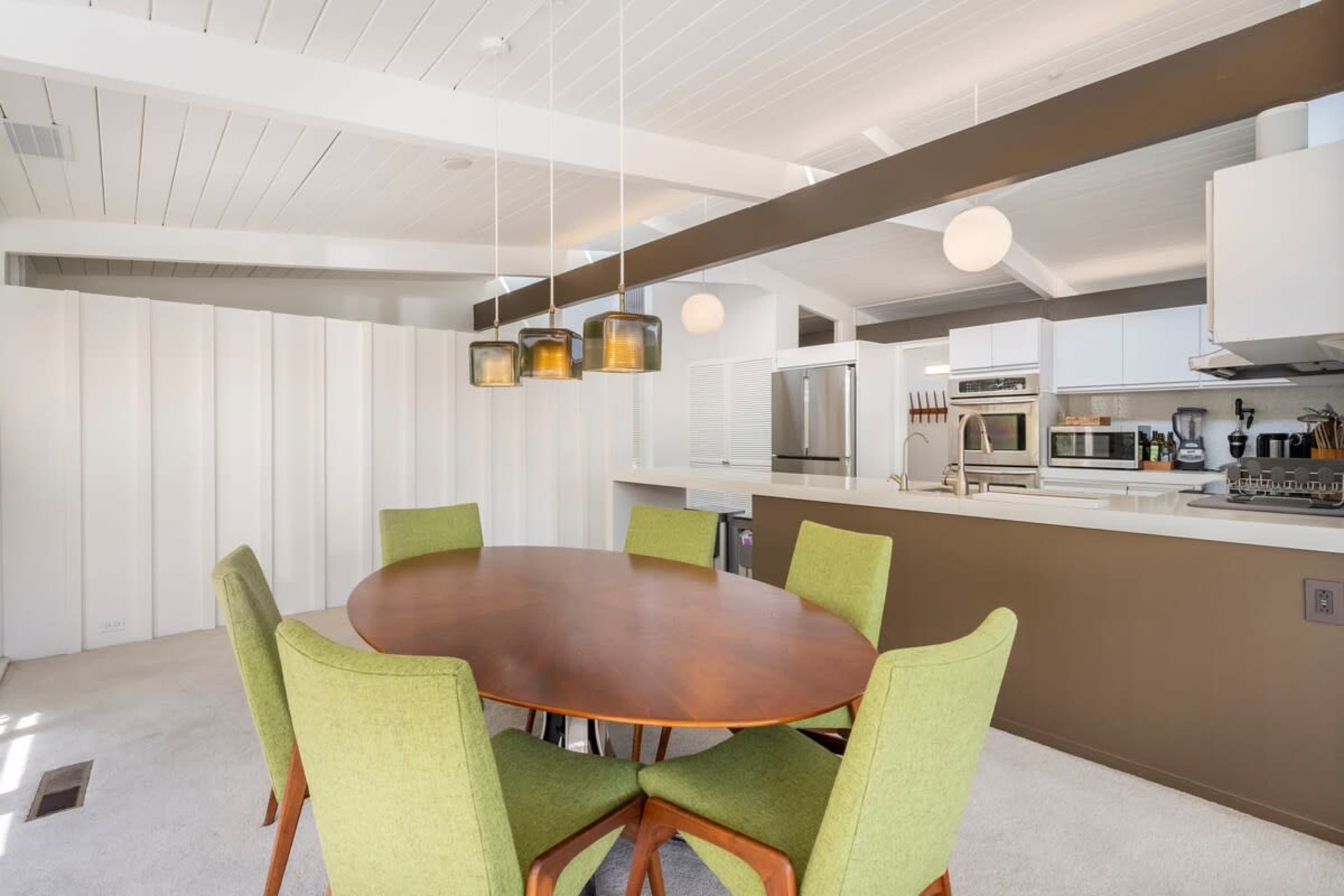 A modern kitchen and dining area features a circular wooden table surrounded by green chairs, with overhead pendant lights and a kitchen in the background.