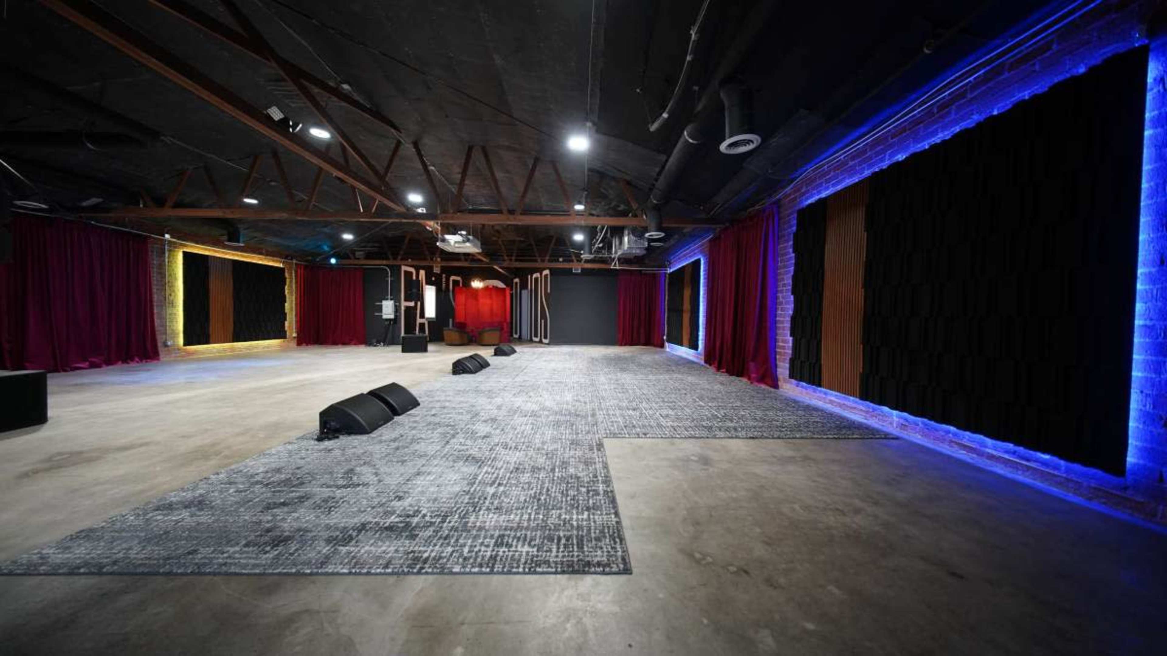 The image shows a spacious event venue featuring a carpeted floor, stage area with microphones, and walls illuminated by colored LED lights.
