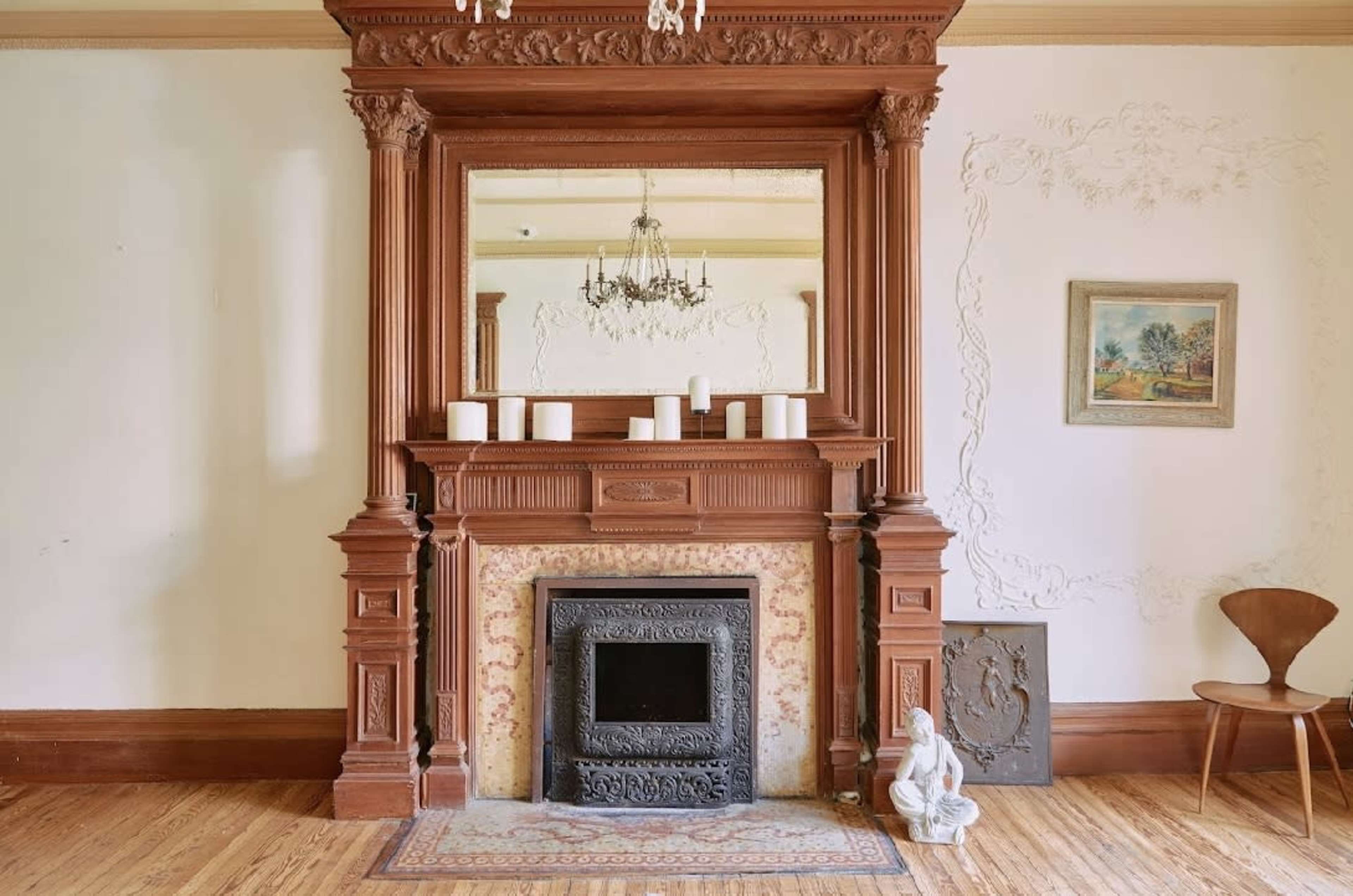 The image features an ornate wooden fireplace mantel with a mirror above it, a black fireplace insert, candles on the mantel, and a small statue beside a wooden chair against a light-colored wall.