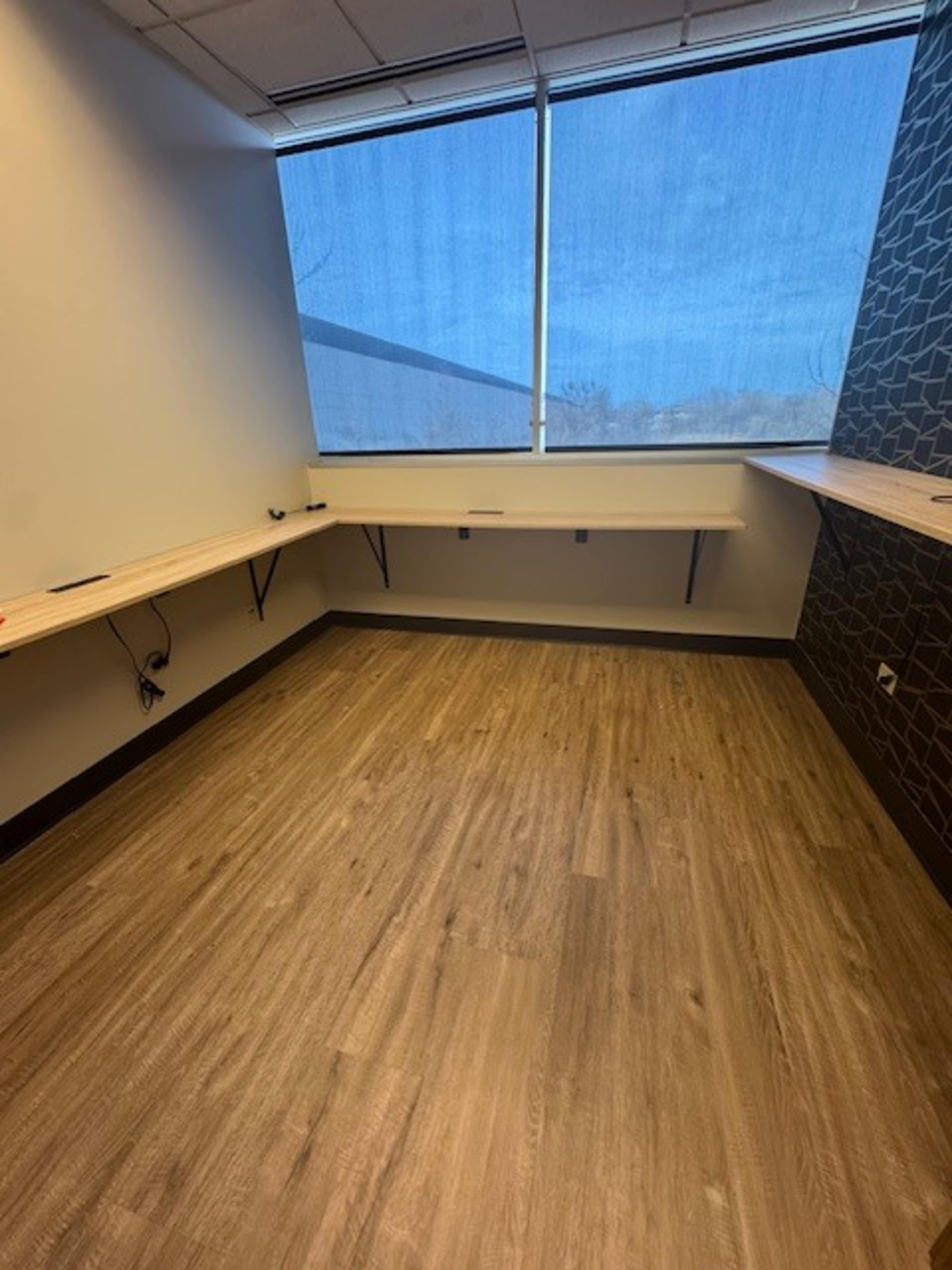 The image shows a sparse office space with wooden countertops along two walls and large windows with blue curtains.