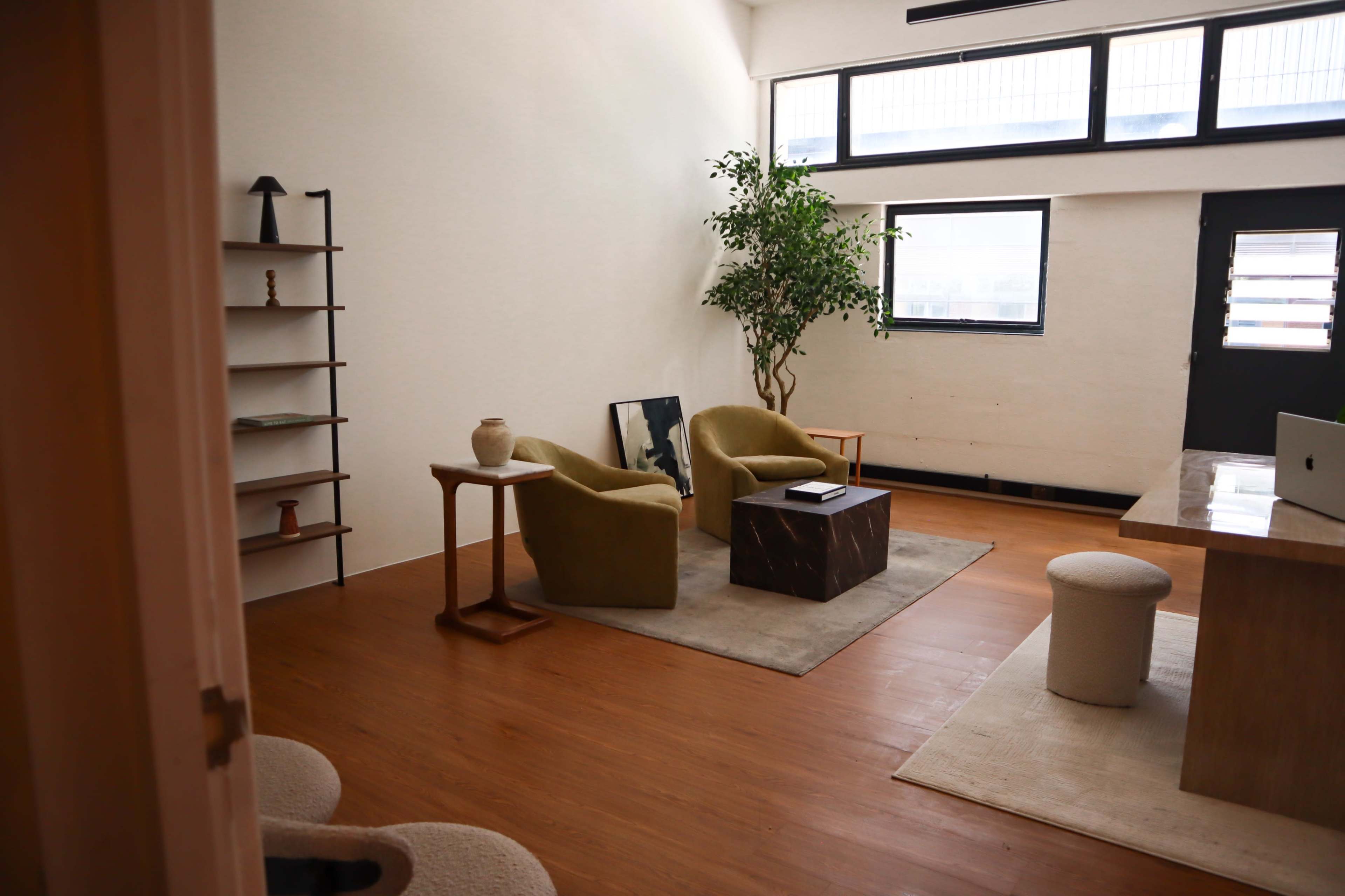 The image shows a modern living space with two green chairs, a small table, a potted plant, and large windows that allow natural light to enter.