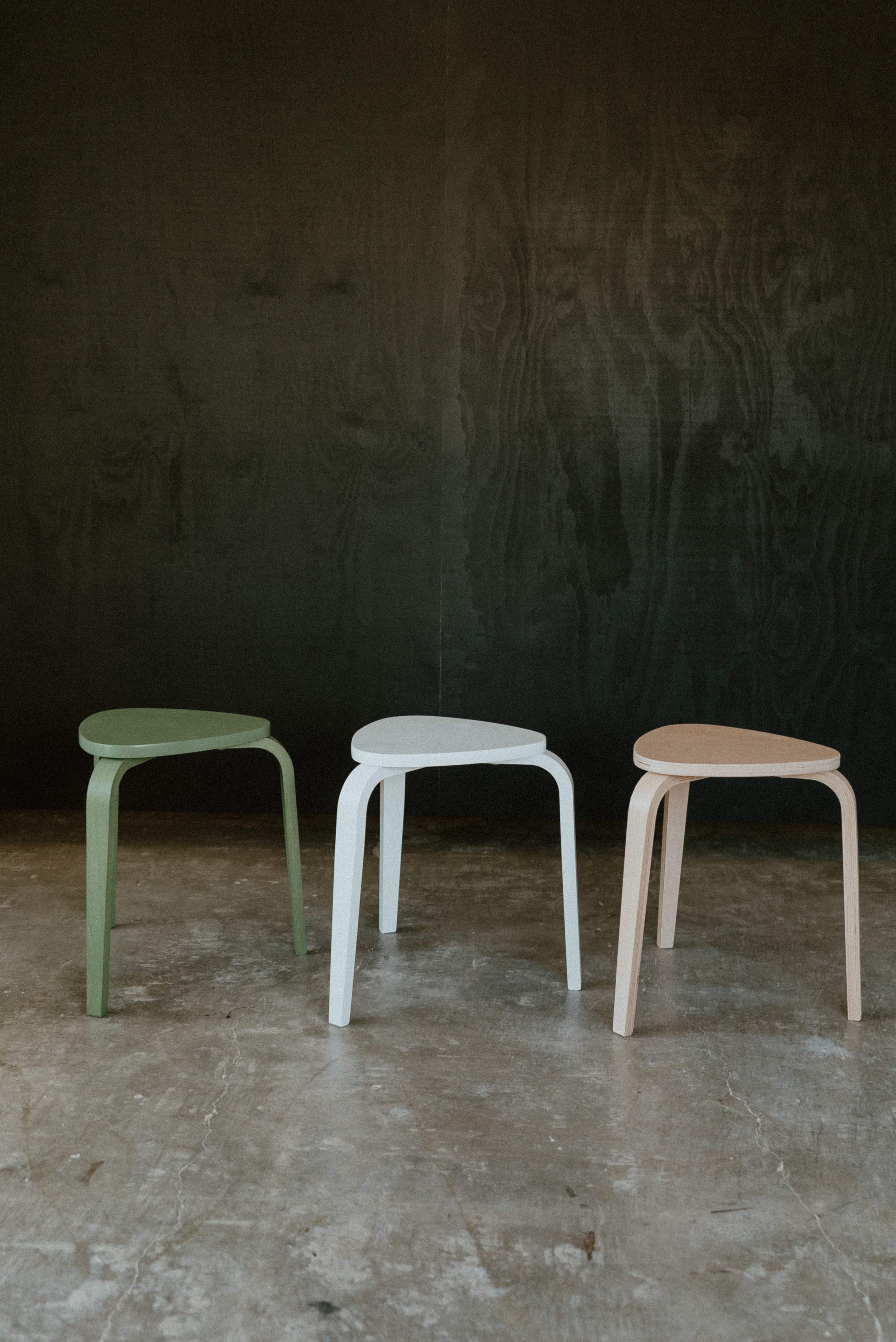 Three minimalist stools in green, white, and light brown are arranged in front of a dark textured backdrop.