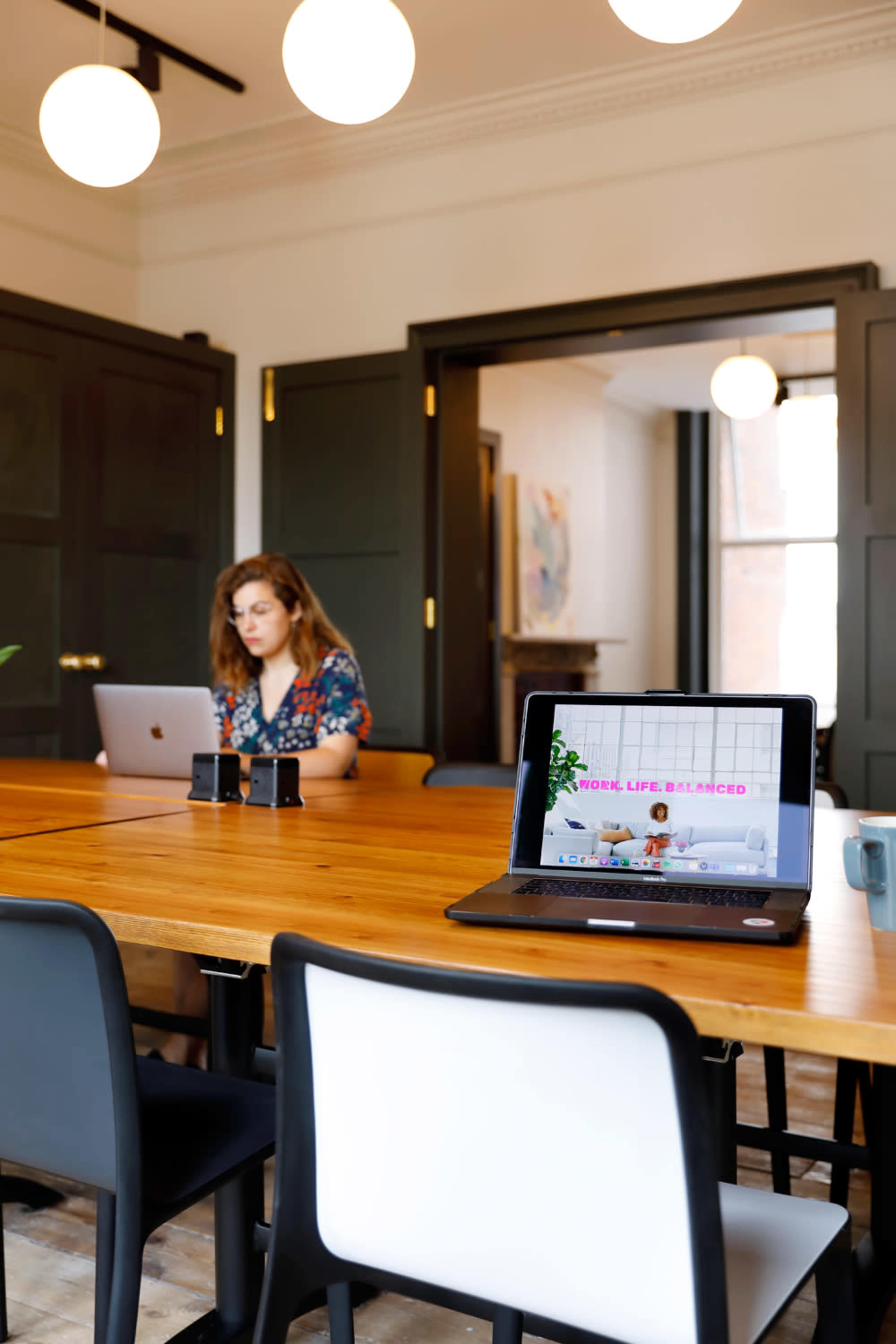 A person works on a laptop at a wooden table, while another individual sits at a nearby laptop in a modern conference room.