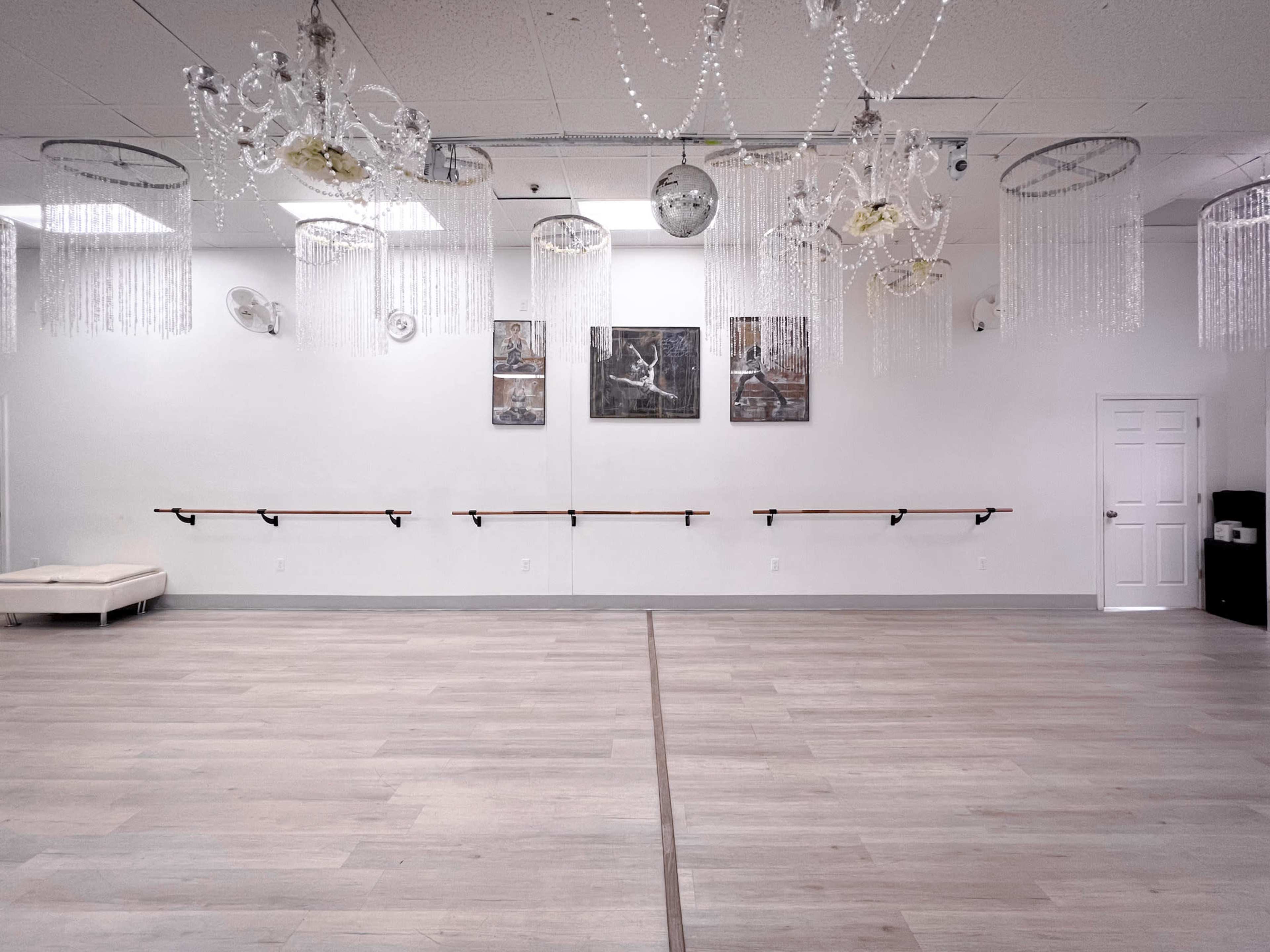 The image shows a large dance studio with wooden flooring, mirrors on the walls, and decorative chandeliers hanging from the ceiling.