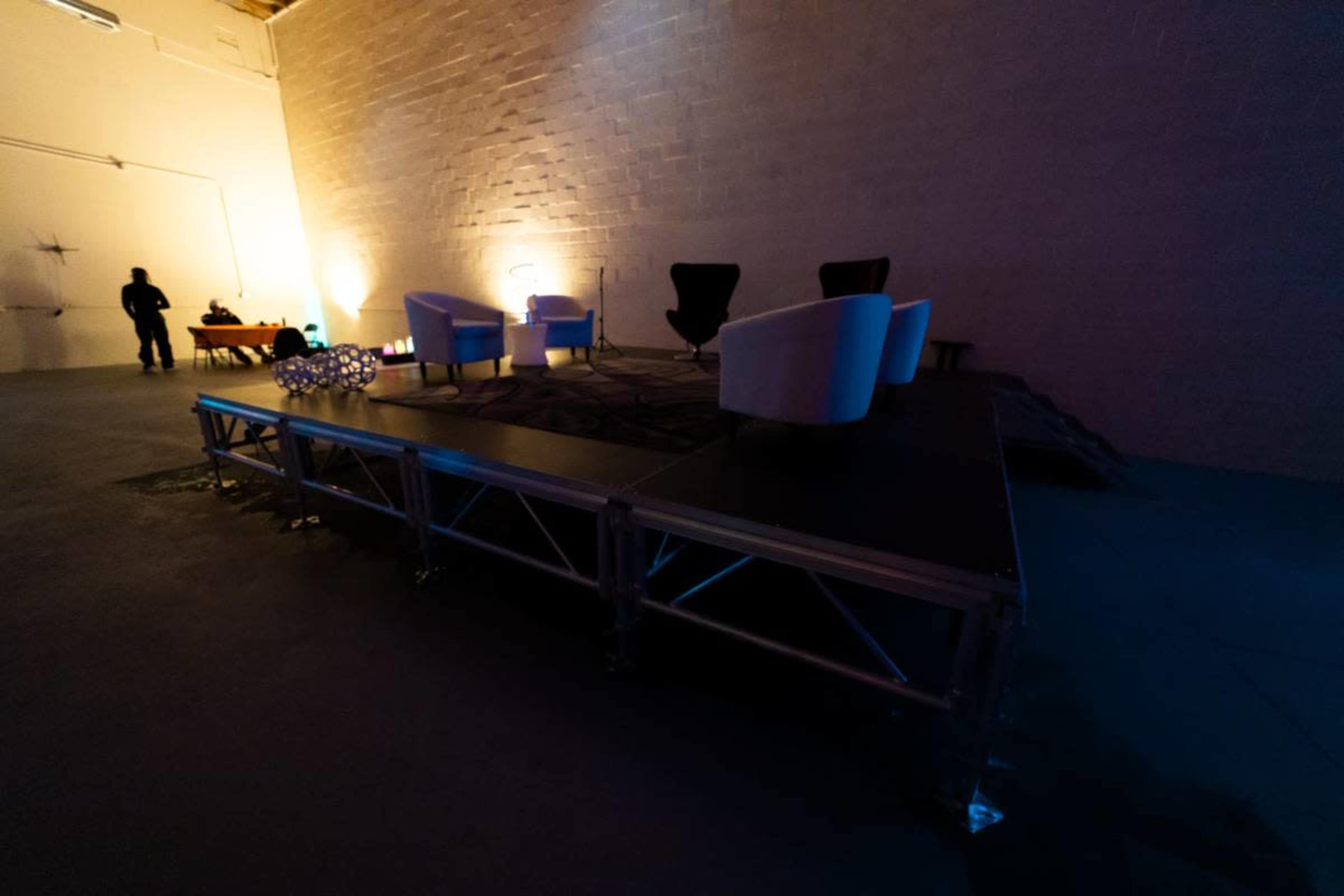 A small stage is set up in a warehouse, featuring three chairs and dim lighting, with a silhouetted figure standing in the background.