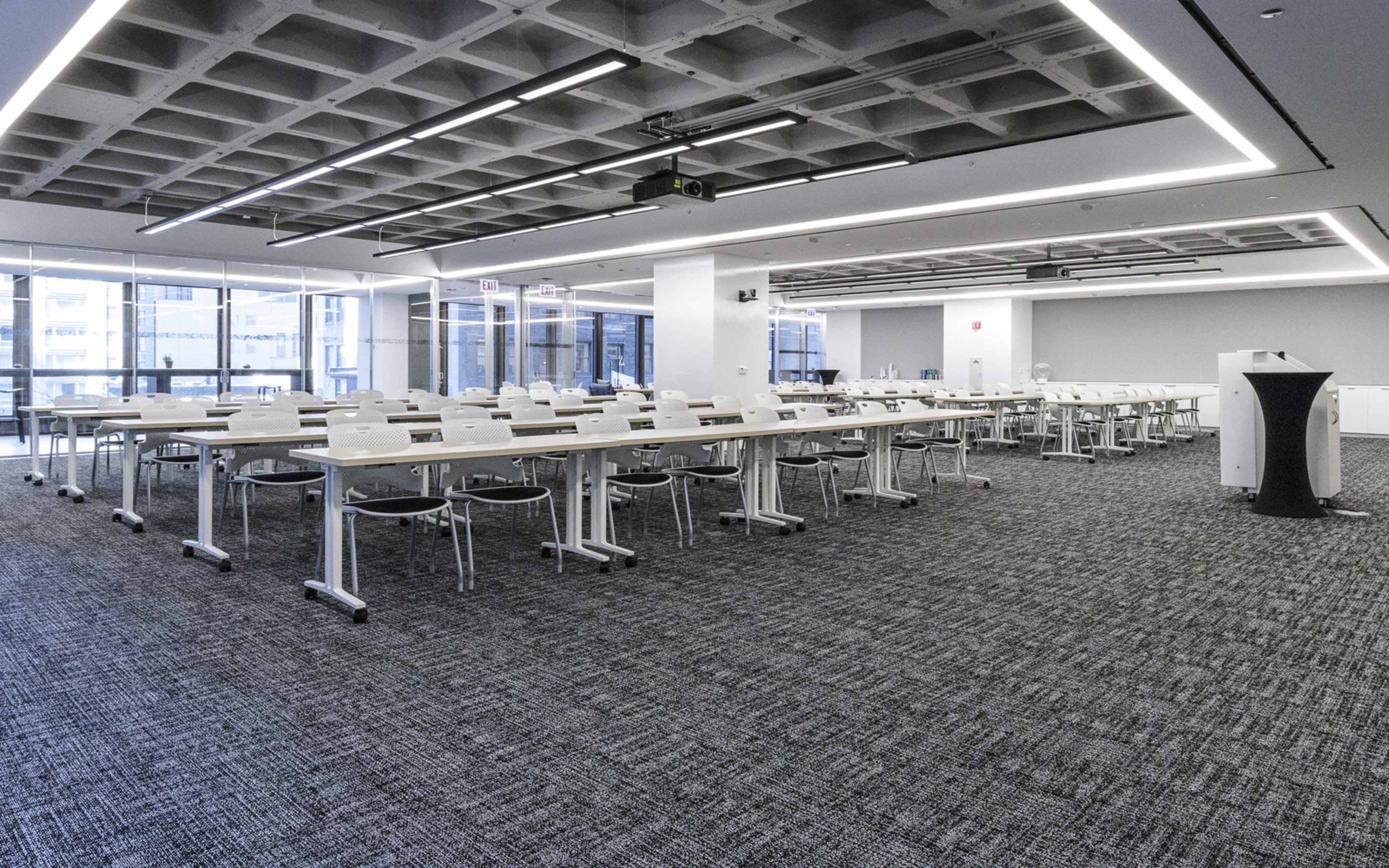 A modern classroom features numerous white tables and chairs arranged in a grid, with large windows allowing natural light and a projector mounted on the ceiling.